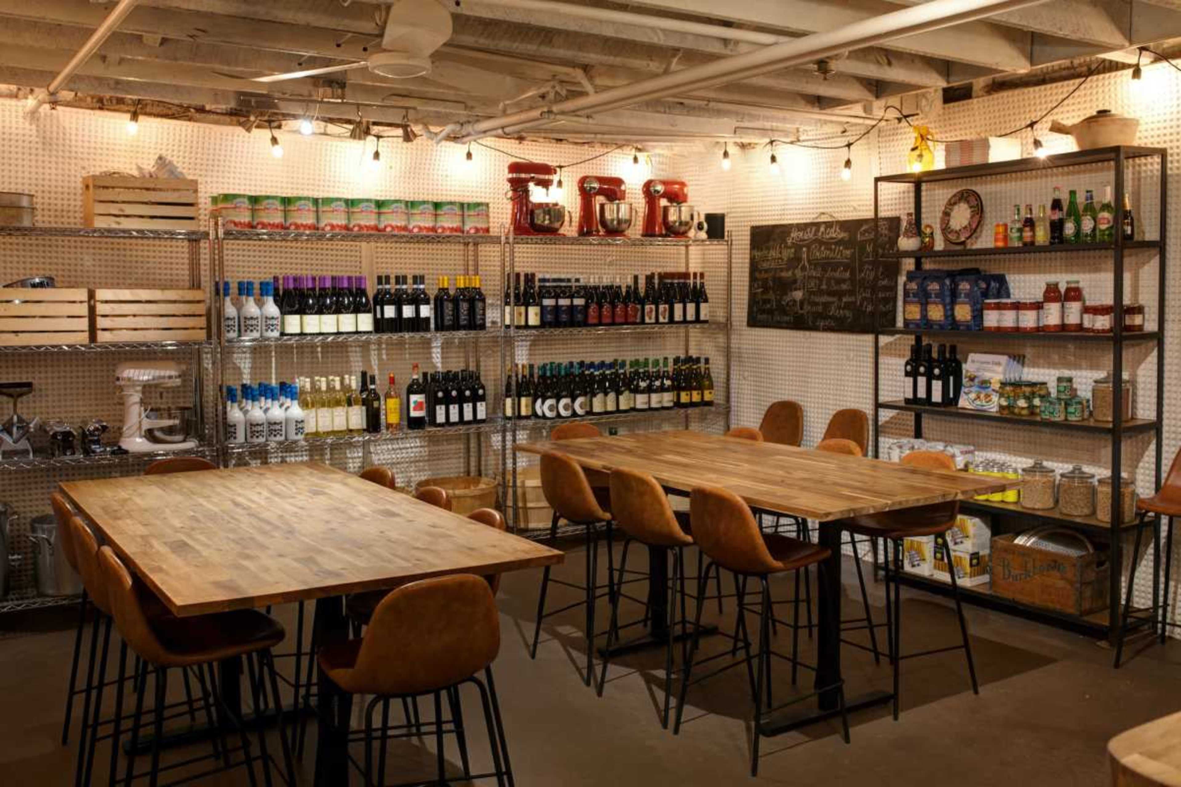 The scene features a well-organized basement space with shelves filled with various bottles, kitchen appliances, and food items, complemented by wooden tables and modern chairs.