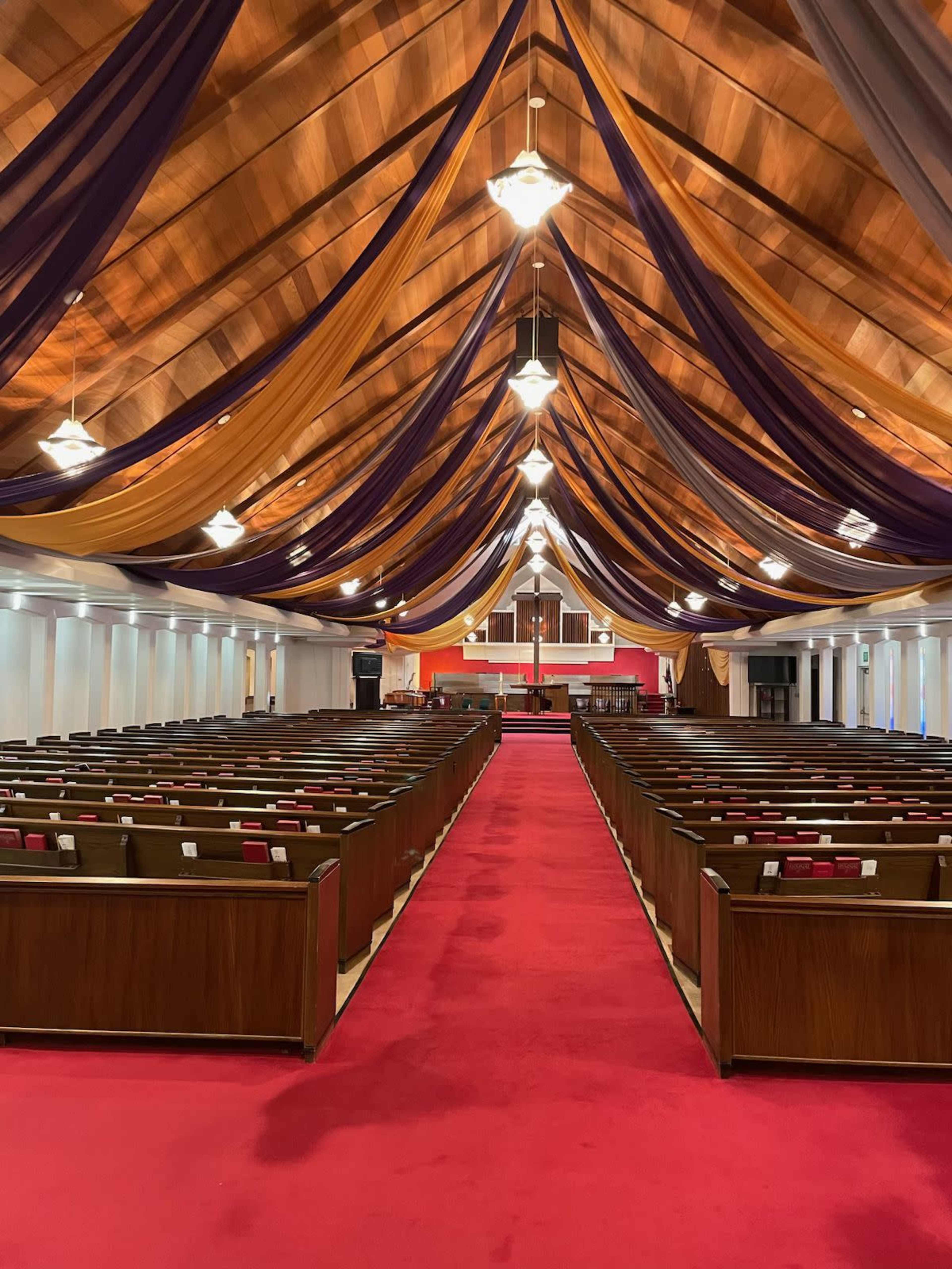 The image shows the interior of a church featuring rows of wooden pews, a red carpeted aisle, and a ceiling draped with fabric in various colors.