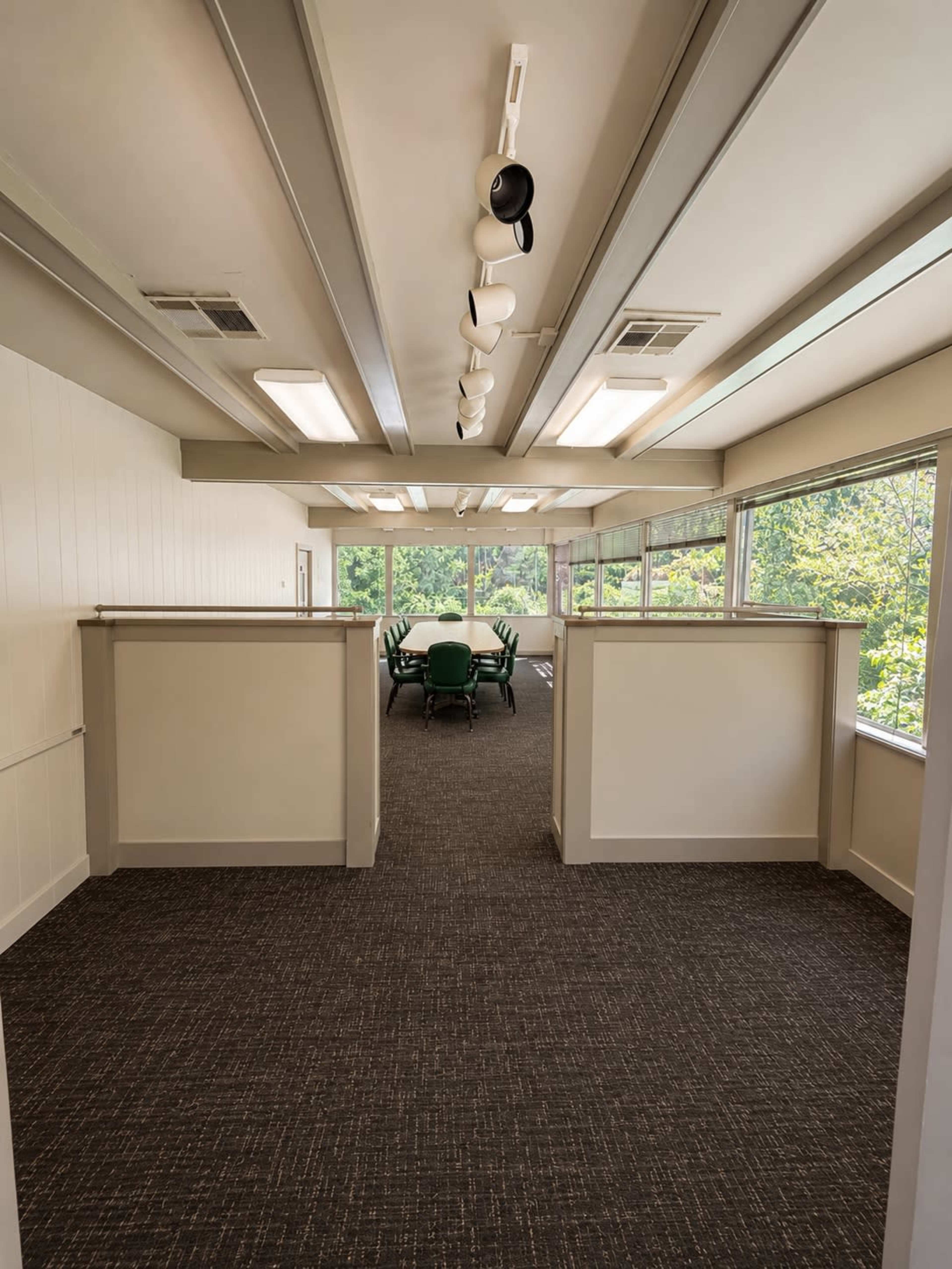 The image shows a bright, modern office space with a long corridor leading to a meeting area, featuring large windows and a green conference table.