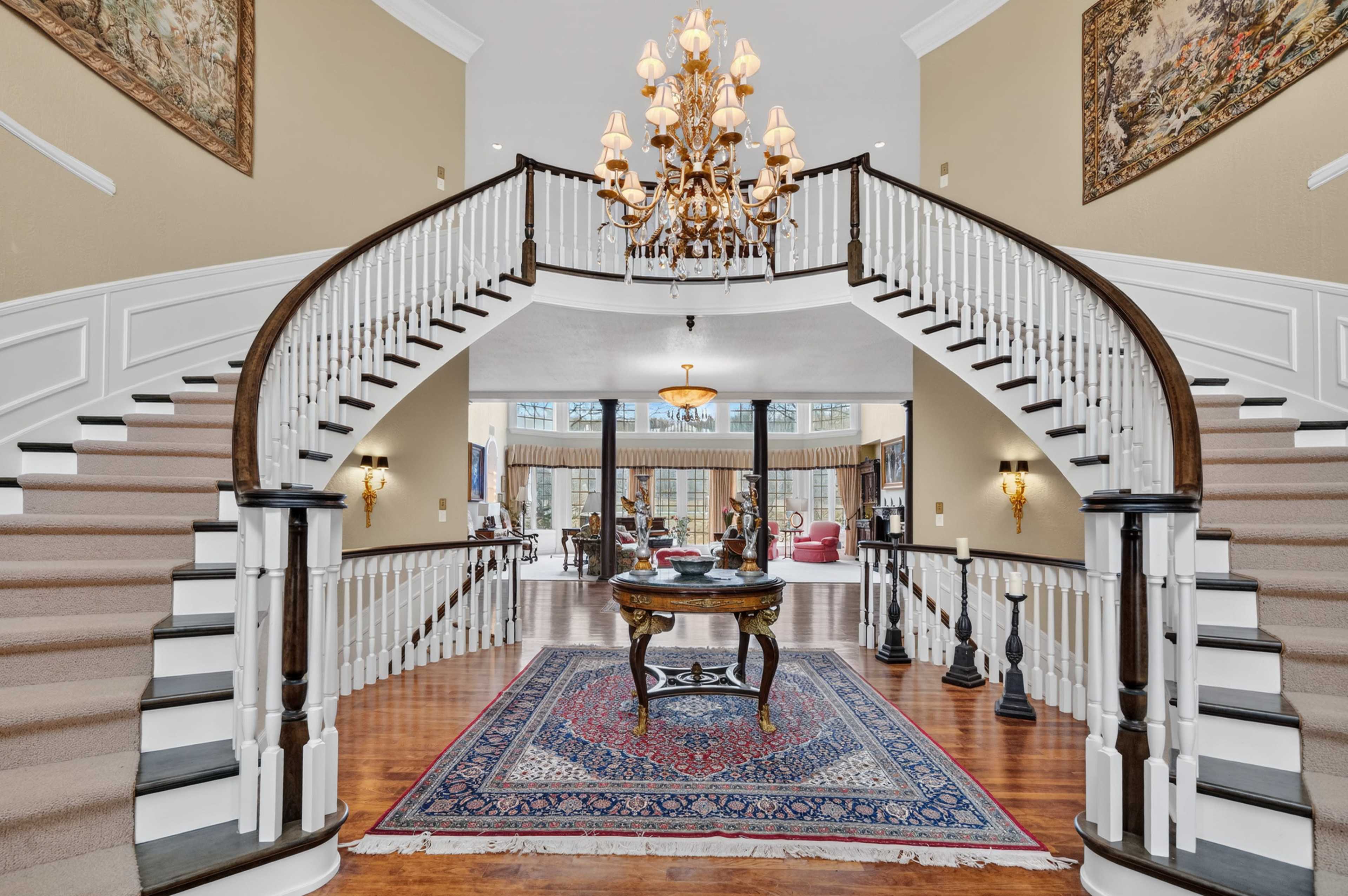 The image depicts a spacious entryway featuring a grand staircase with a chandelier above and a large rug adorned with intricate patterns.