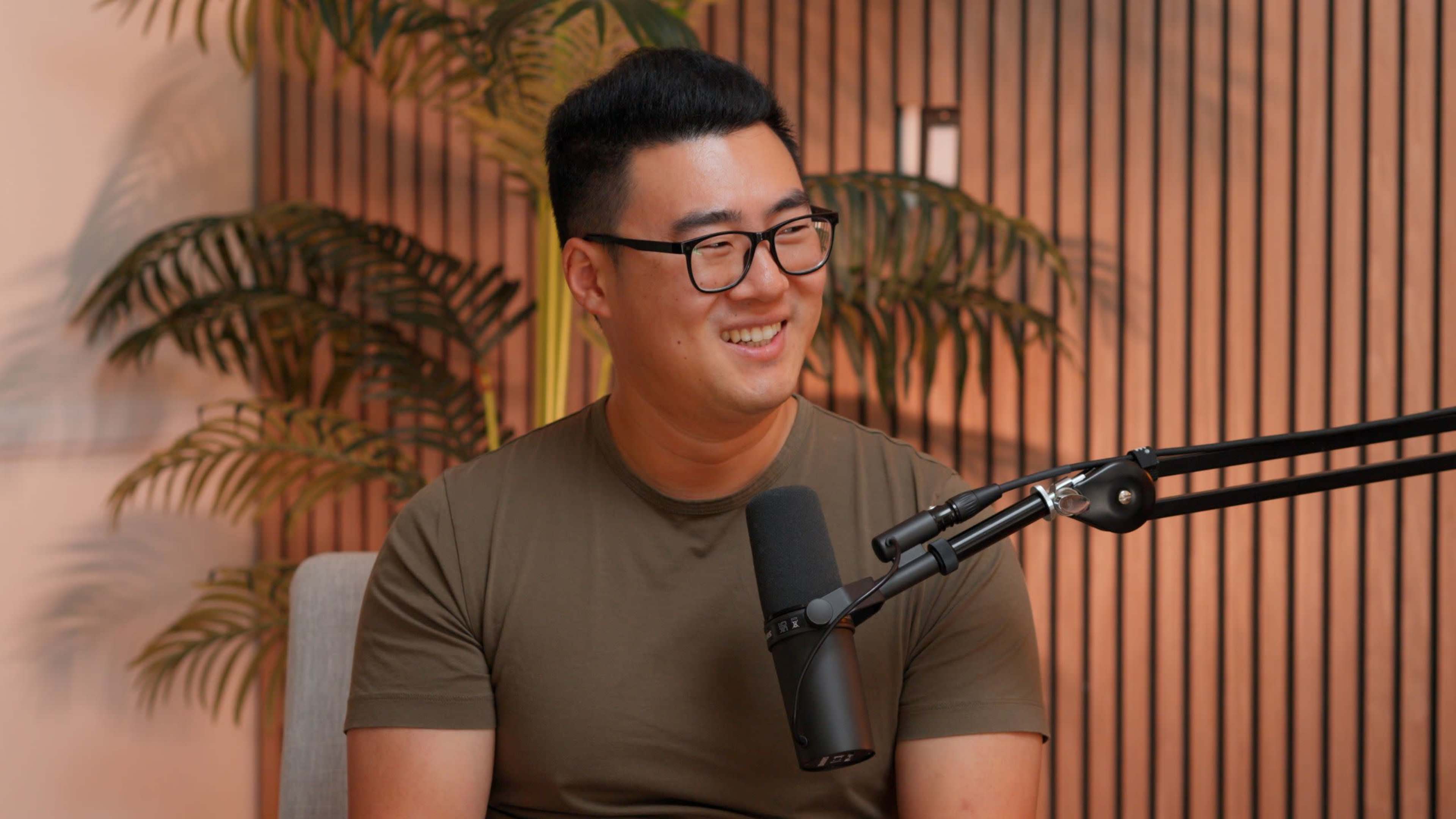 A man wearing glasses and a green shirt sits in front of a microphone against a backdrop of plants and wooden panels.