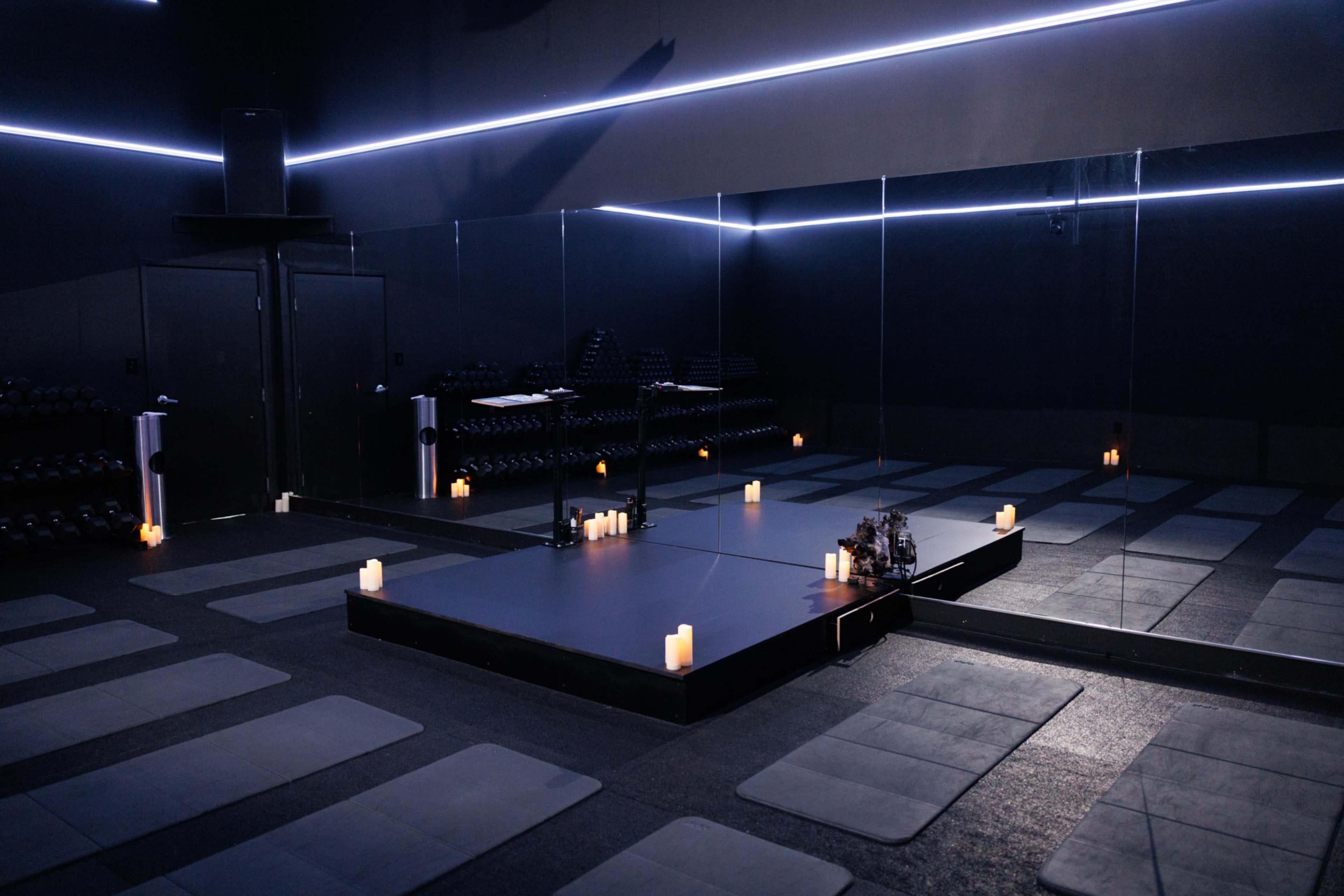 The image shows a dimly lit yoga studio with padded mats arranged on the floor, a mirrored wall, and lit candles placed on a central platform.