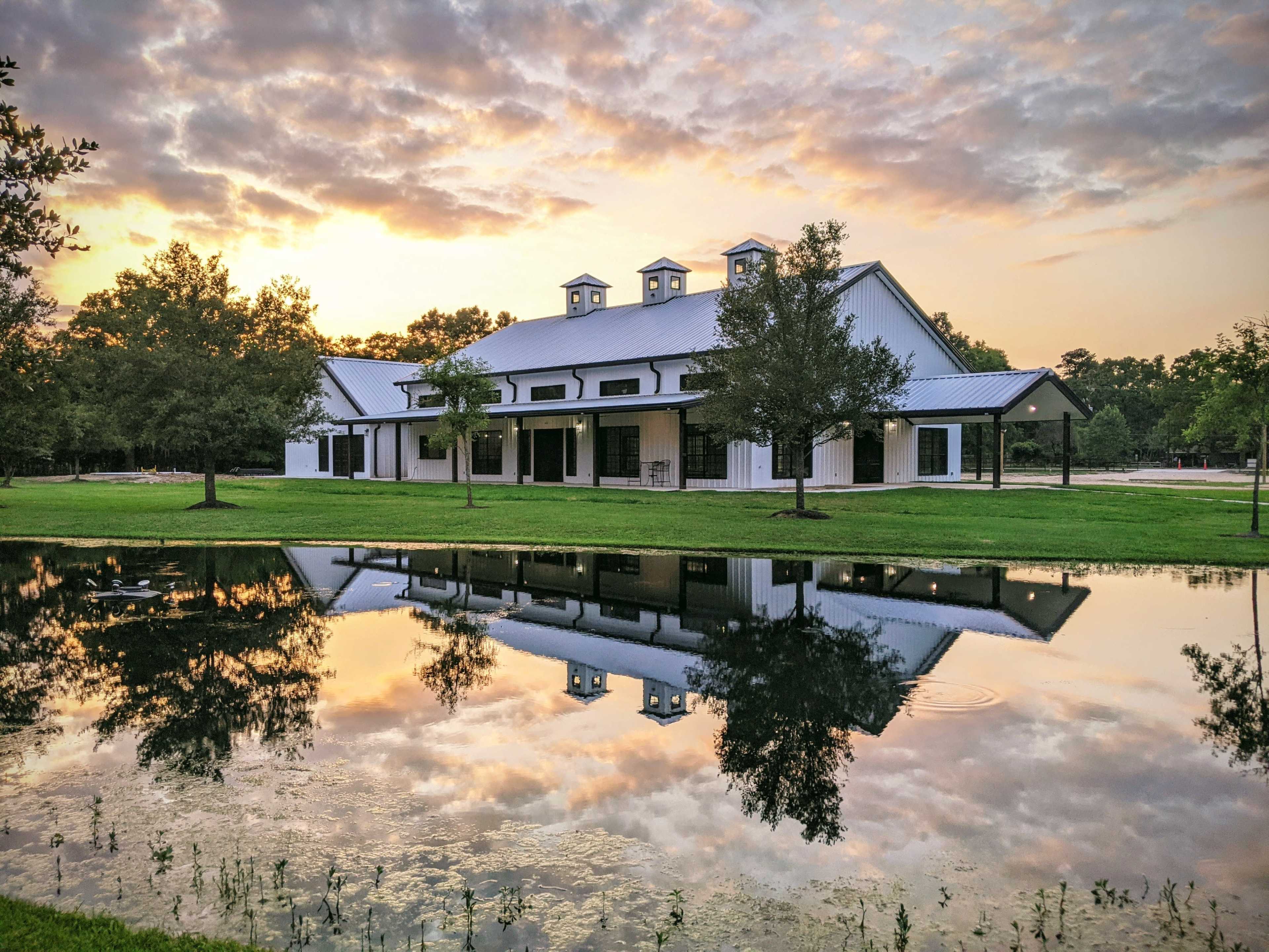 A large white building with a metal roof is reflected in a pond at sunset, surrounded by trees.