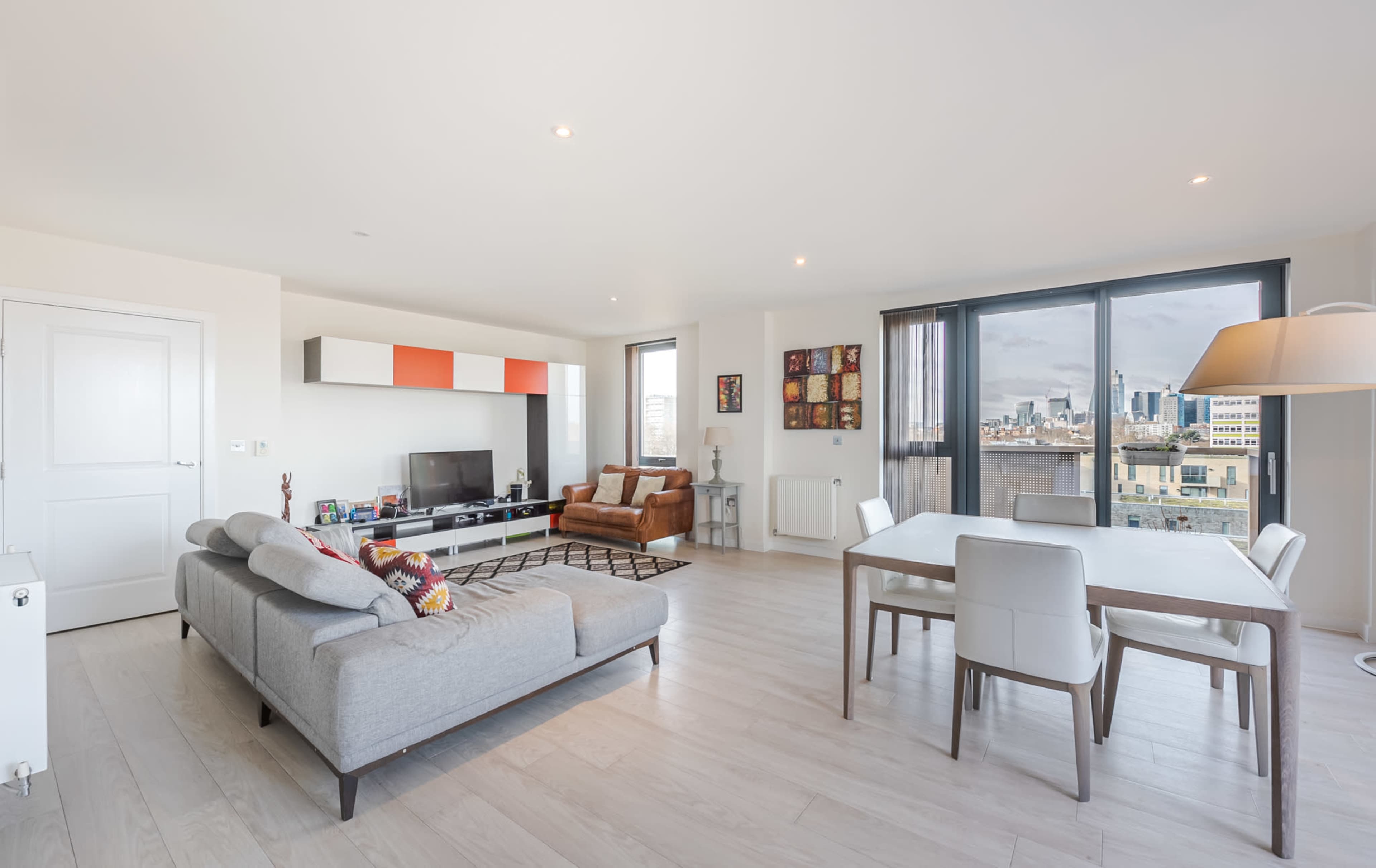 The image shows a modern living room with a sofa, a coffee table, a television unit, a dining table, and large windows overlooking a cityscape.