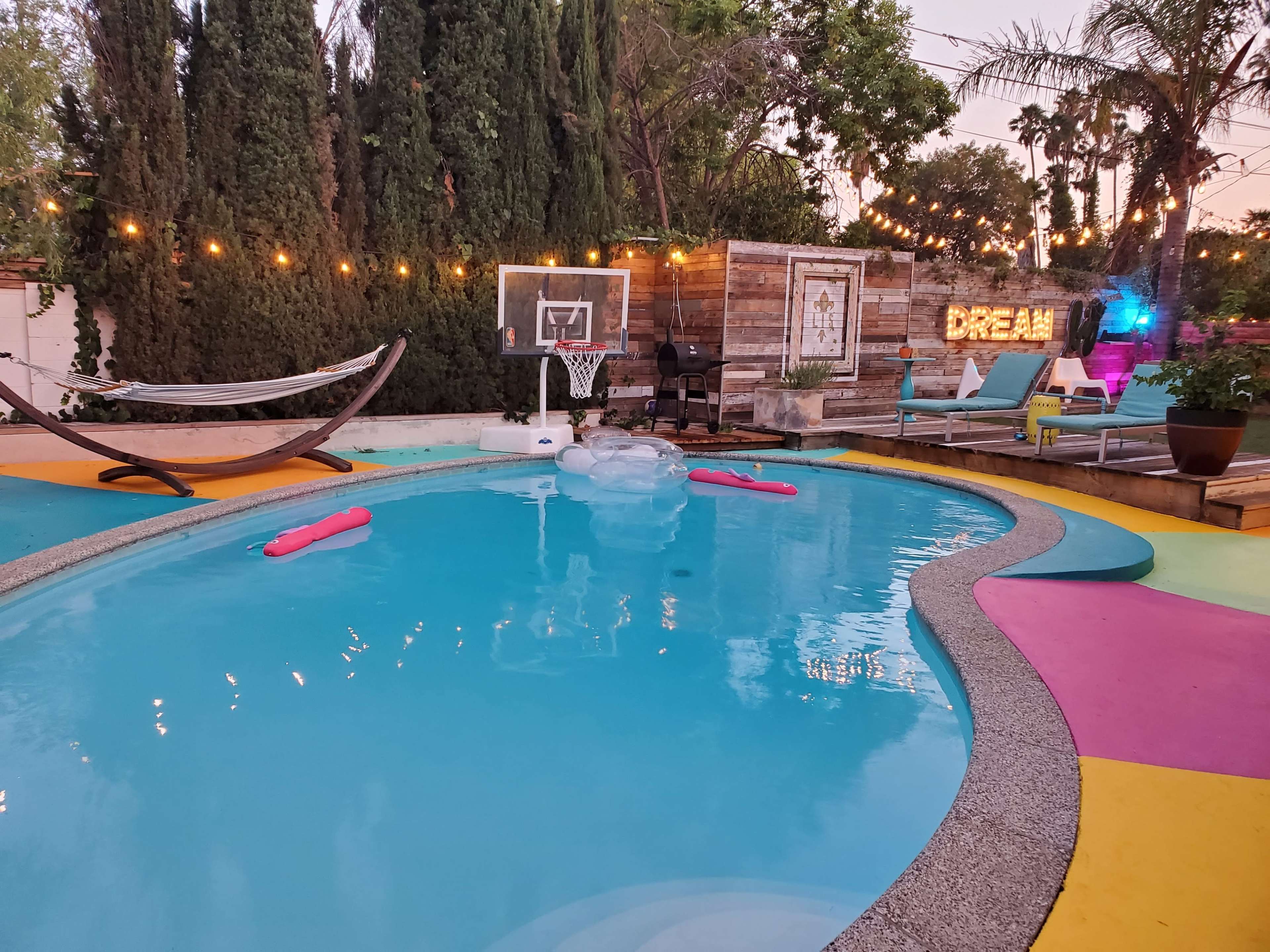 The scene features a swimming pool surrounded by colorful decking, a basketball hoop, a hammock, and illuminated decorations in a landscaped backyard.
