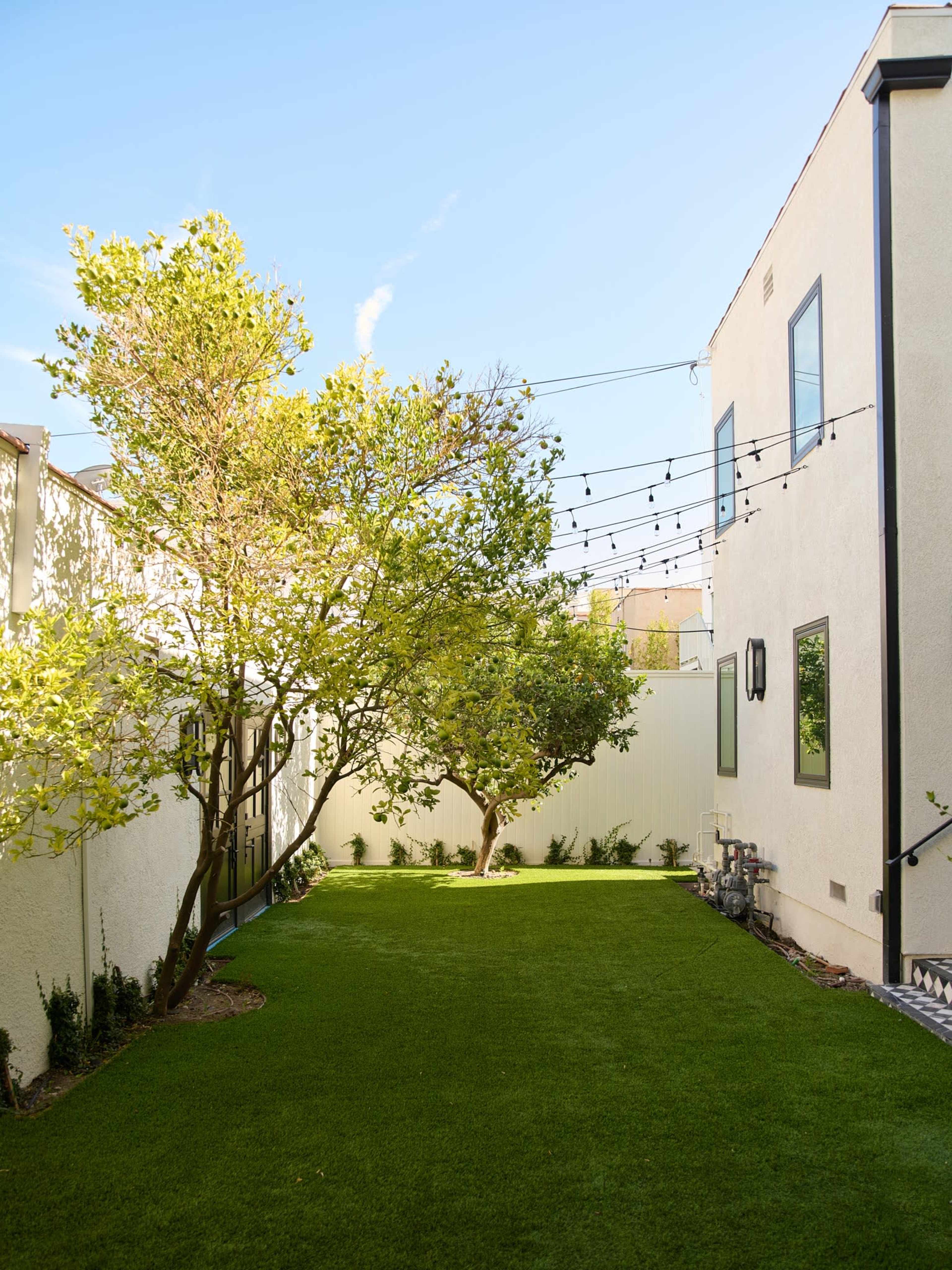 The image shows a narrow backyard with neatly maintained artificial grass, two trees, and string lights overhead.
