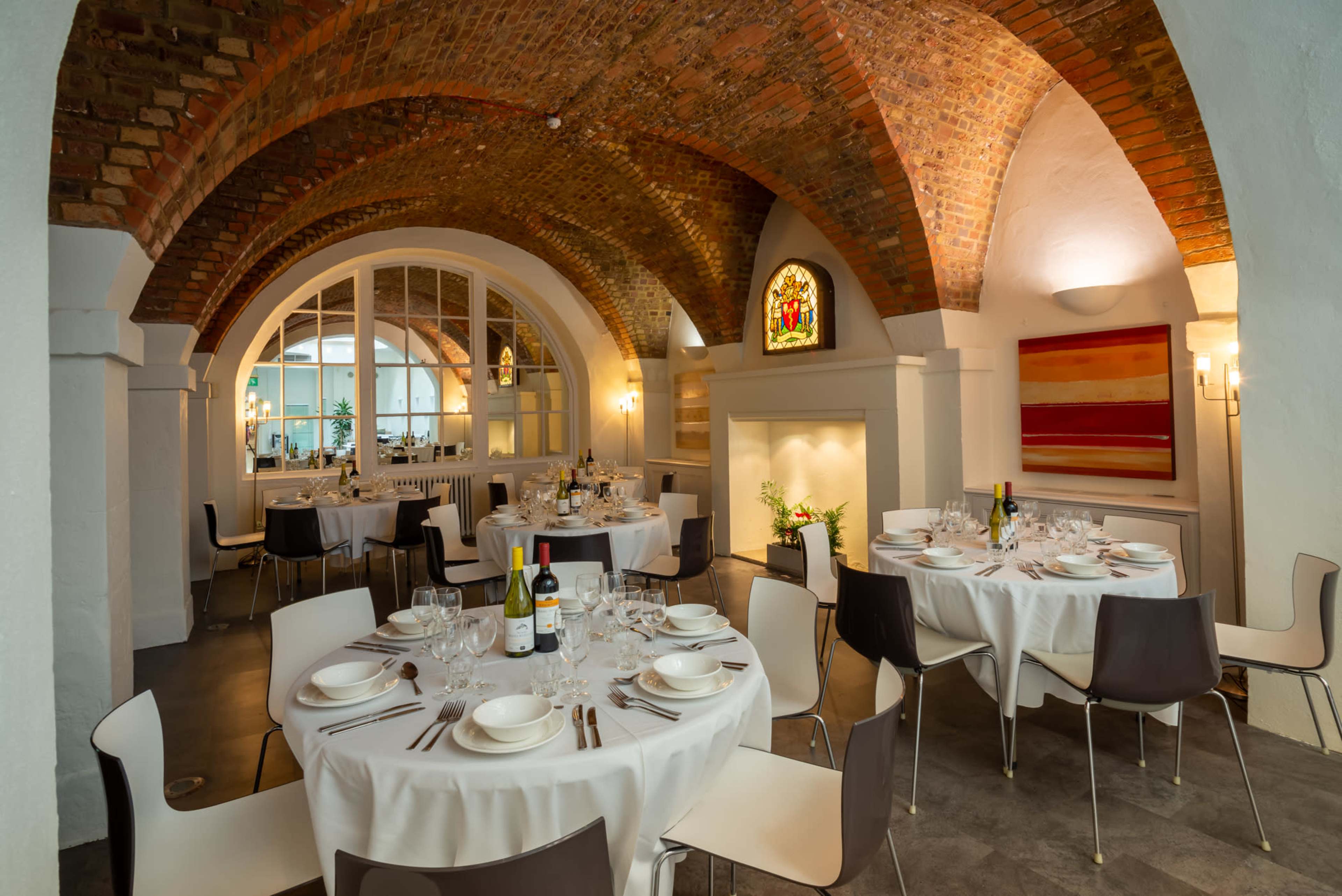 A dining area in a restaurant with arched brick ceilings, round tables set with white tablecloths, and large windows letting in light.