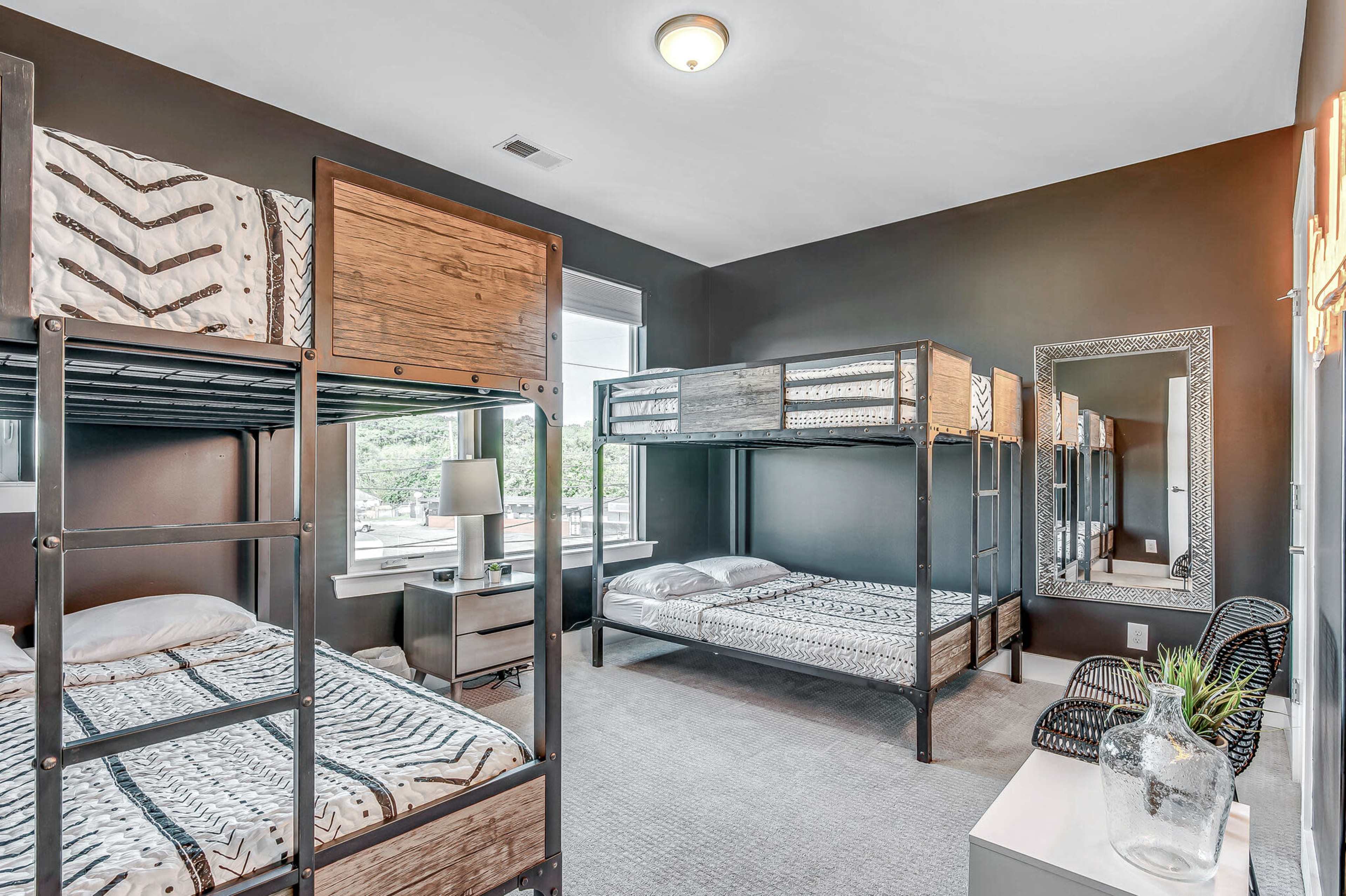 The image shows a bedroom furnished with two metal bunk beds, a nightstand with a lamp, and a large mirror, all set against dark-colored walls.