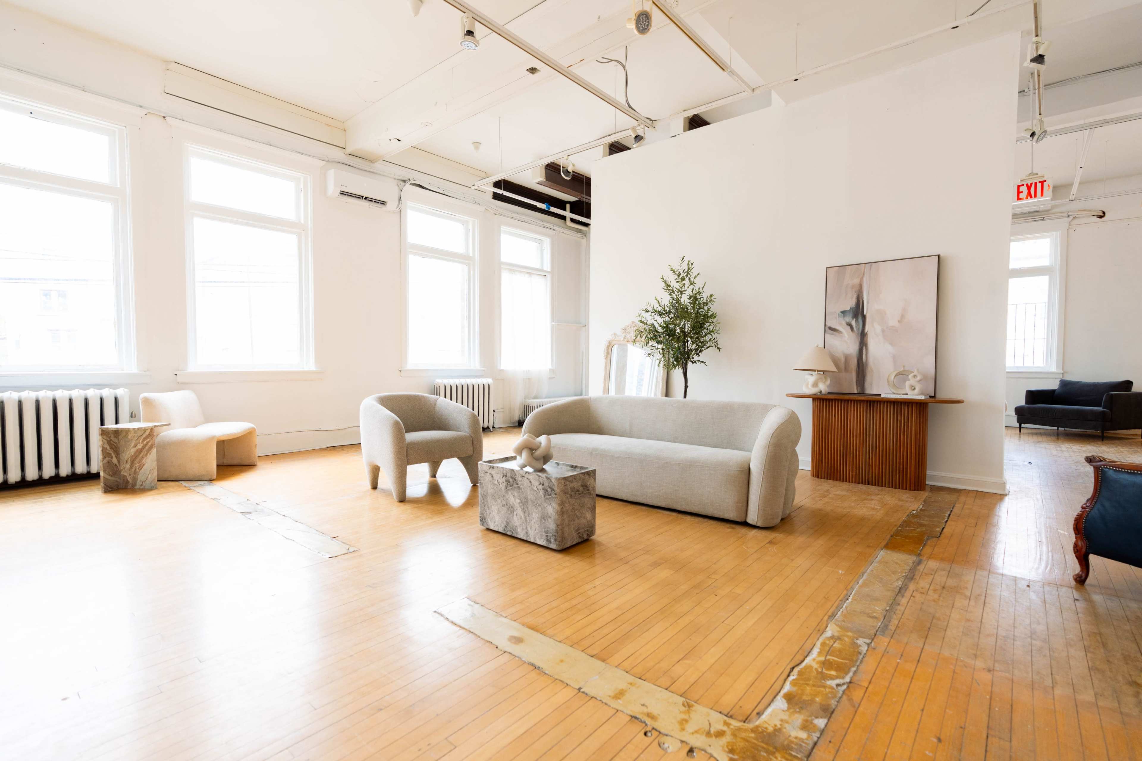 The image depicts a spacious, well-lit room featuring a curved sofa, two armchairs, a small table made of stone, a minimalist art piece, and large windows.