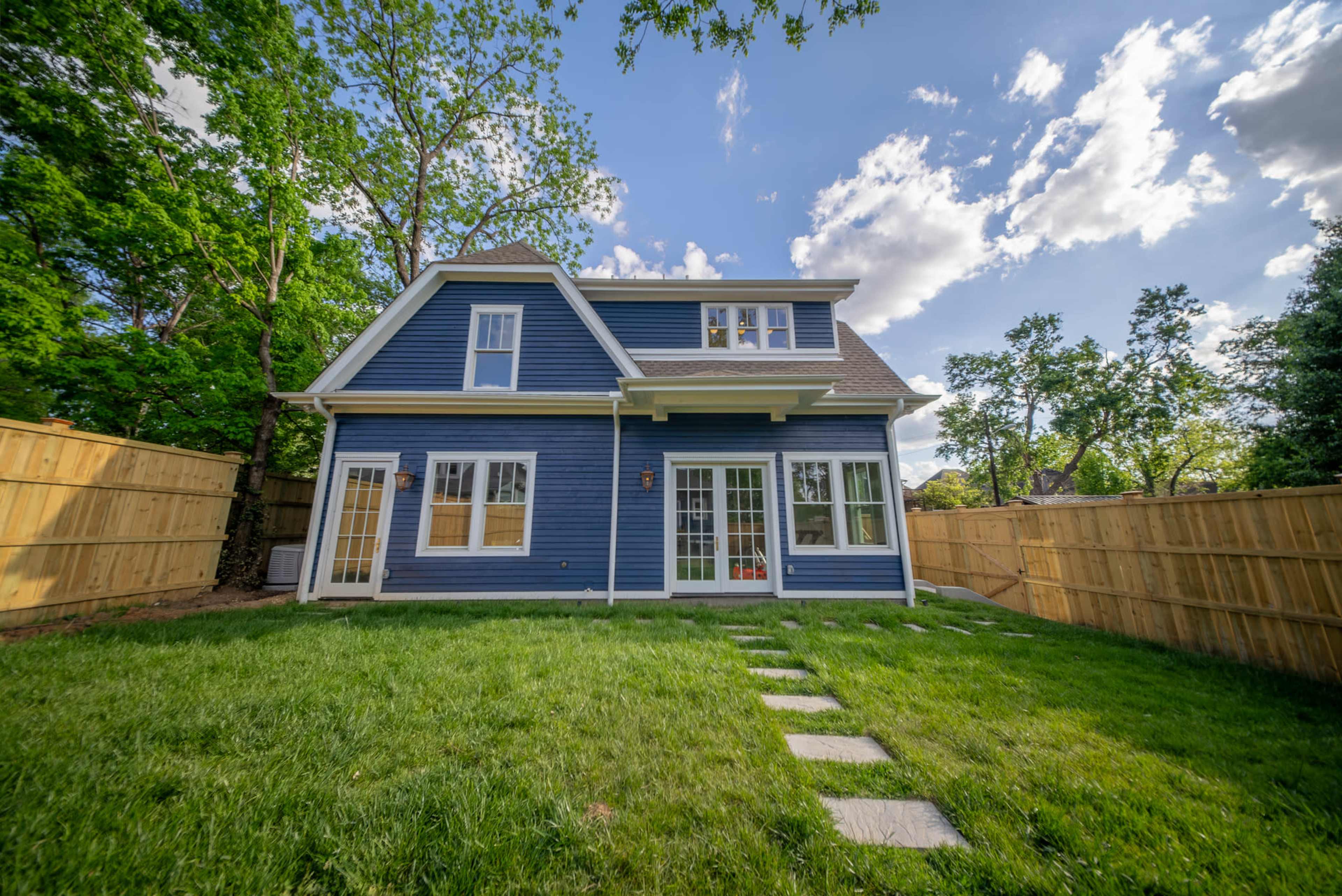 A blue two-story house with a sloped roof stands in a green backyard surrounded by wooden fences and dotted with stepping stones.