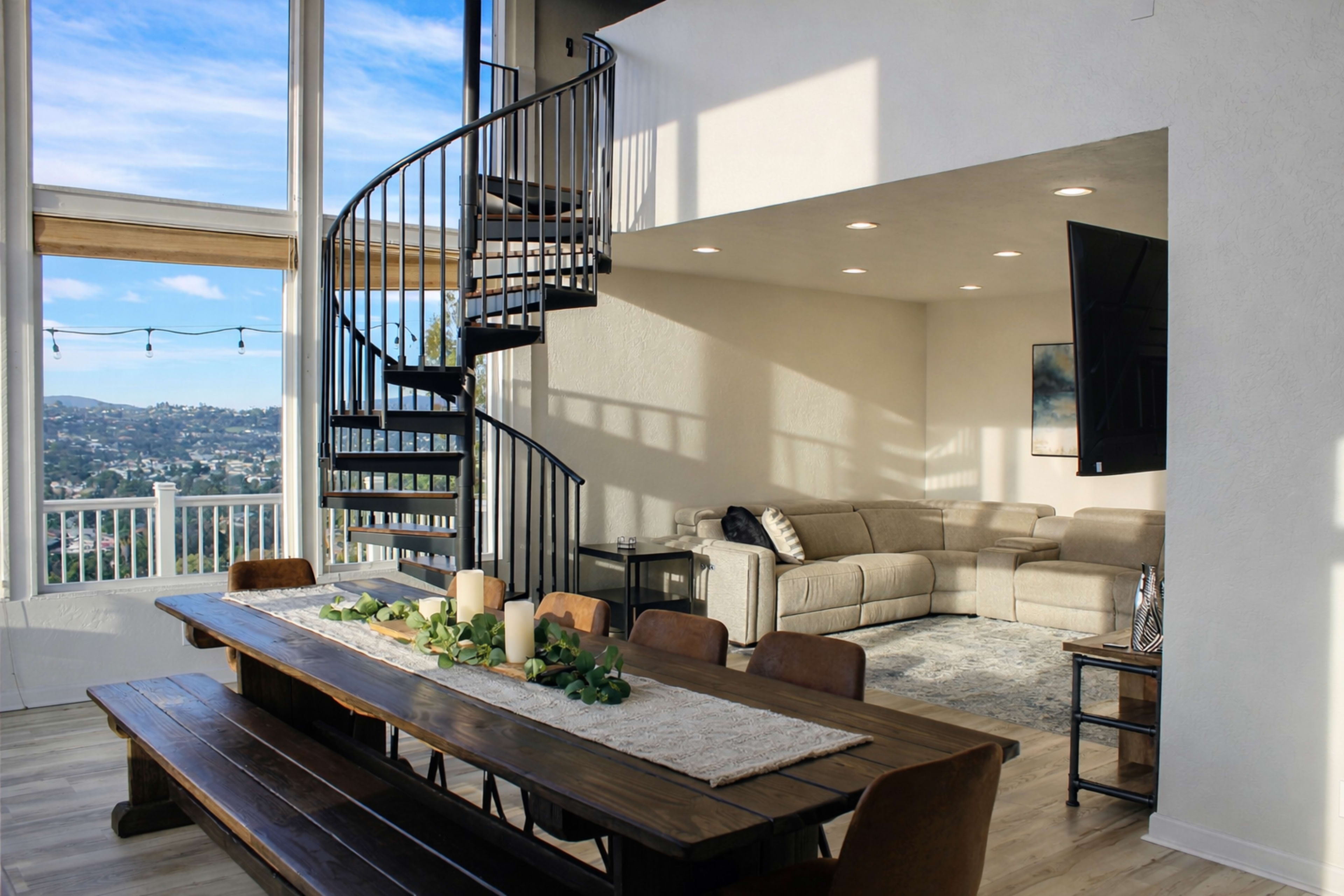 The image shows a modern living space with a spiral staircase, a large dining table adorned with greenery and candles, and a cozy seating area with a sectional couch.