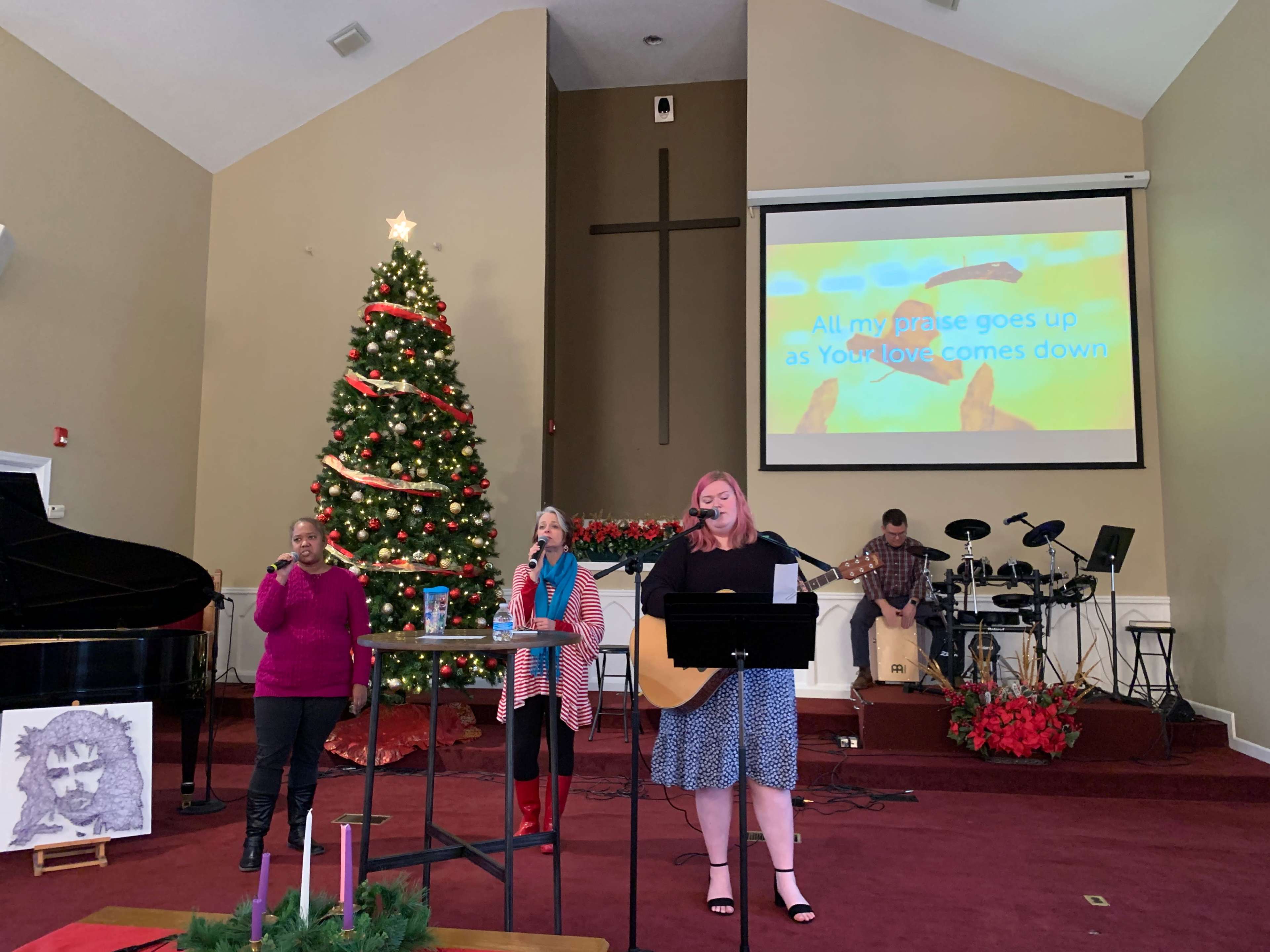 A group of musicians performs in front of a decorated Christmas tree in a church setting, with a projected screen displaying lyrics in the background.