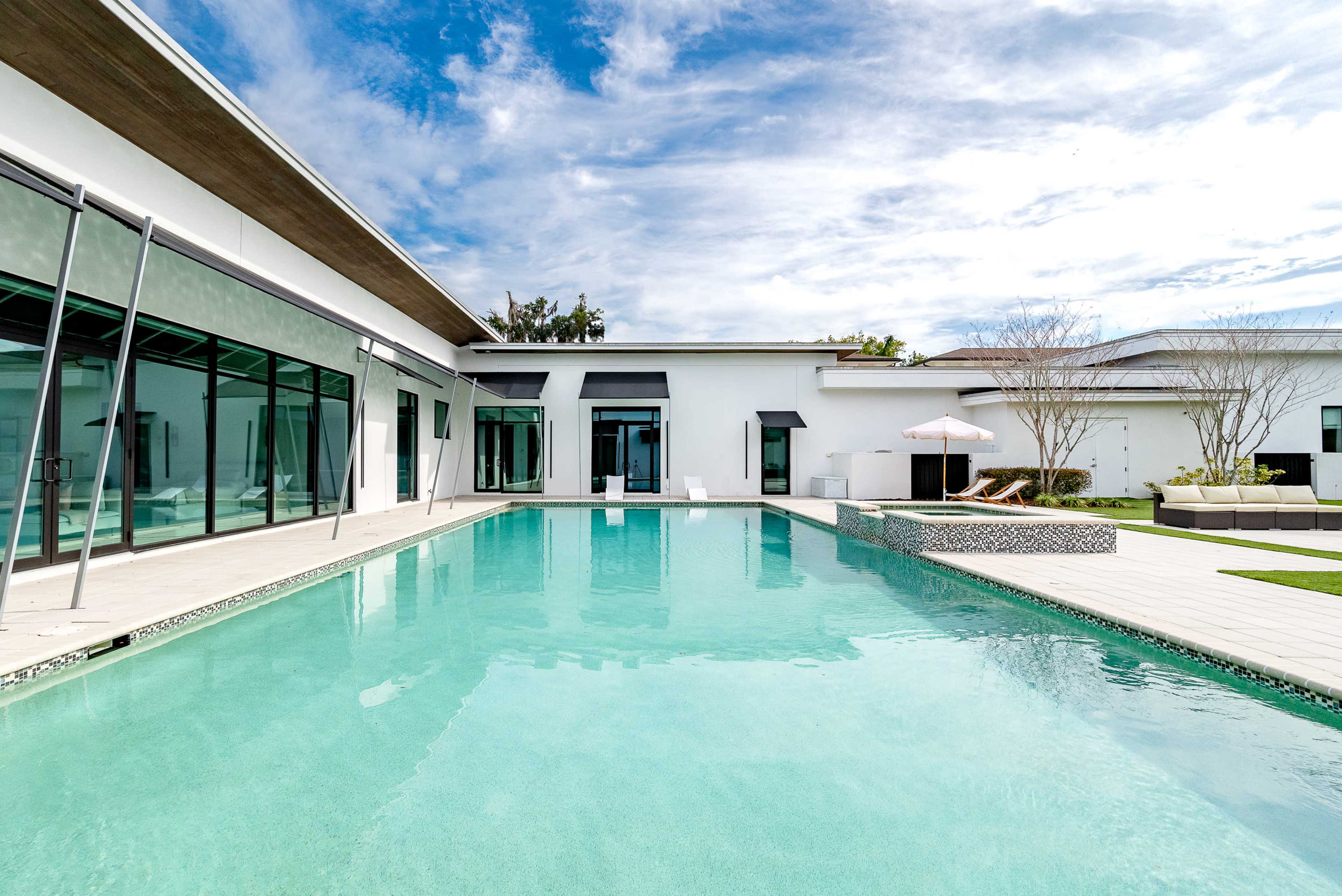 The image shows a modern house with large glass windows surrounding a clear swimming pool.