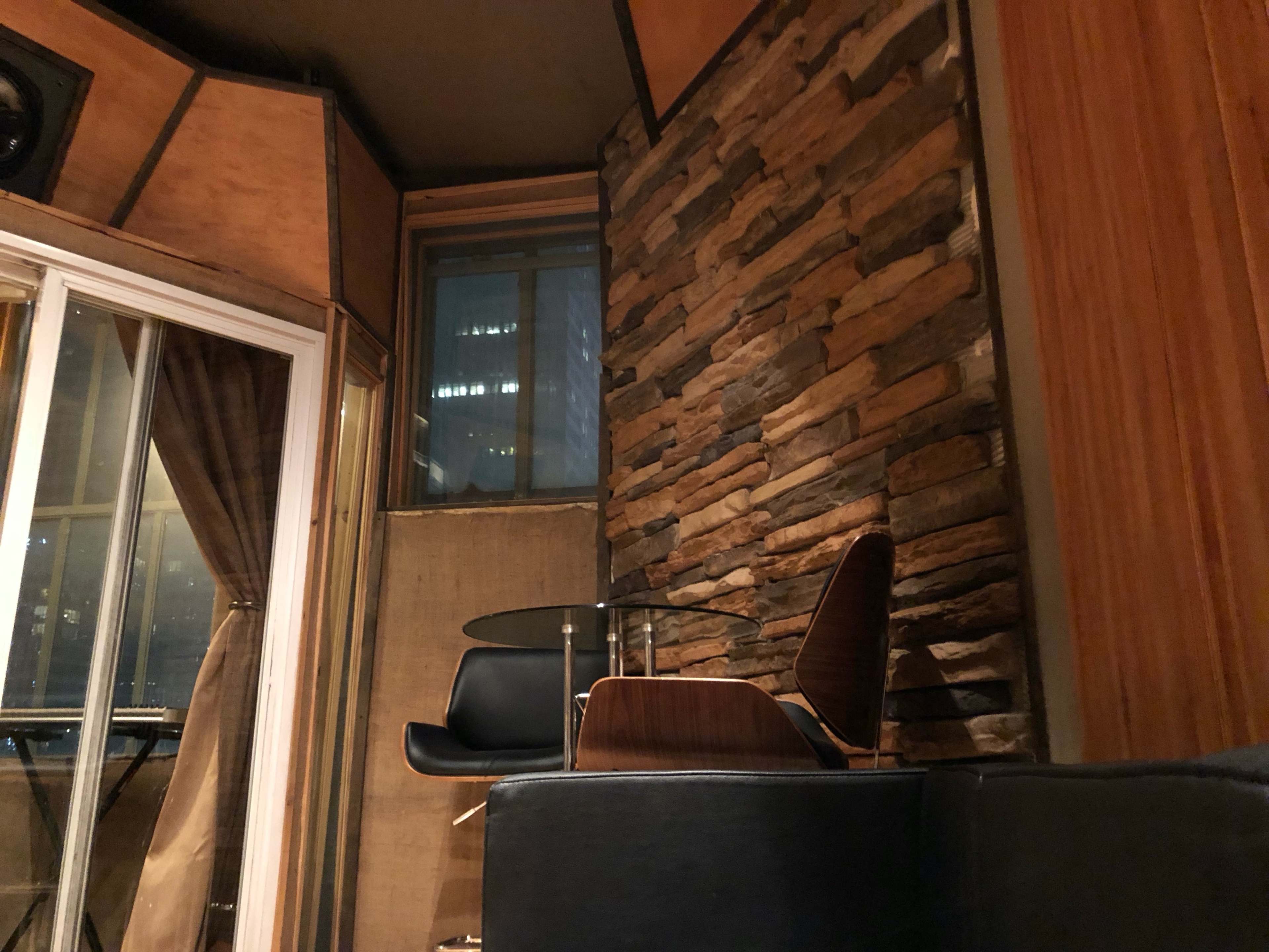 The image shows a corner of a room with a stone feature wall, a glass table, and a chair, with large windows revealing a skyline outside.