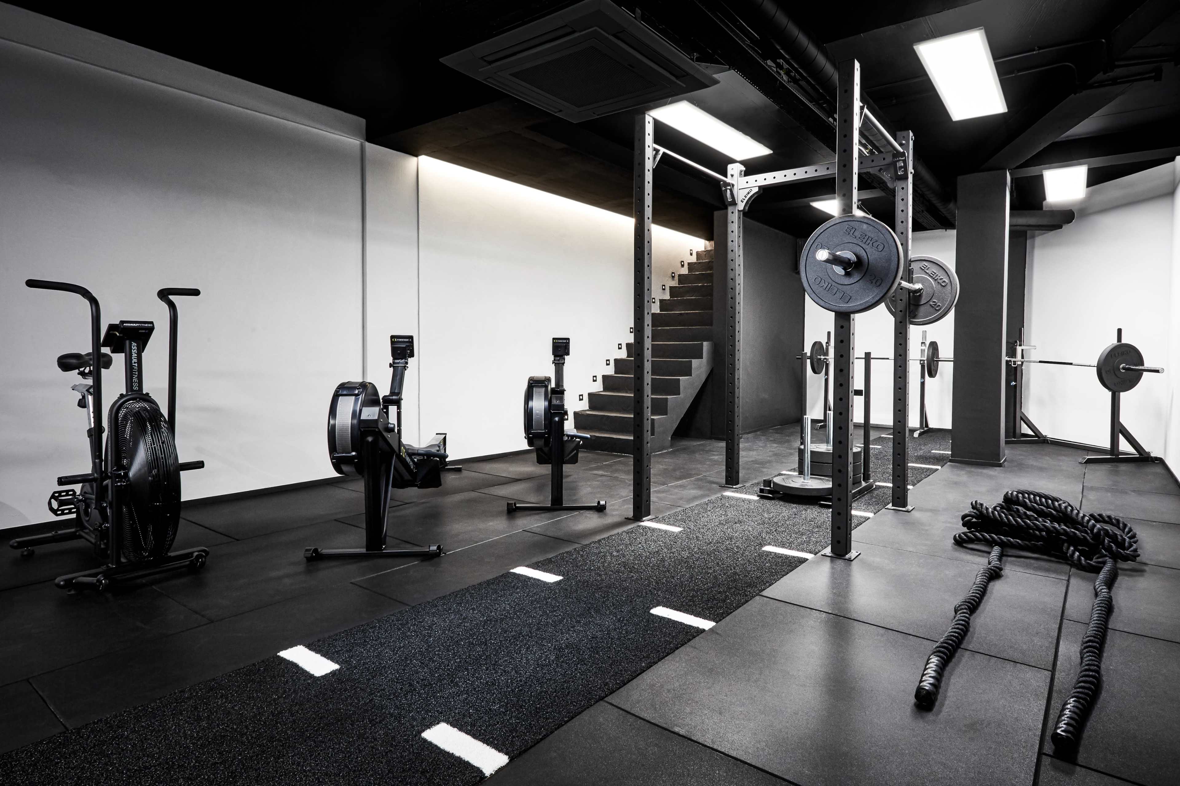 A modern gym with exercise equipment, including rowing machines, weightlifting bars, and battle ropes, set against a minimalist black and white backdrop.