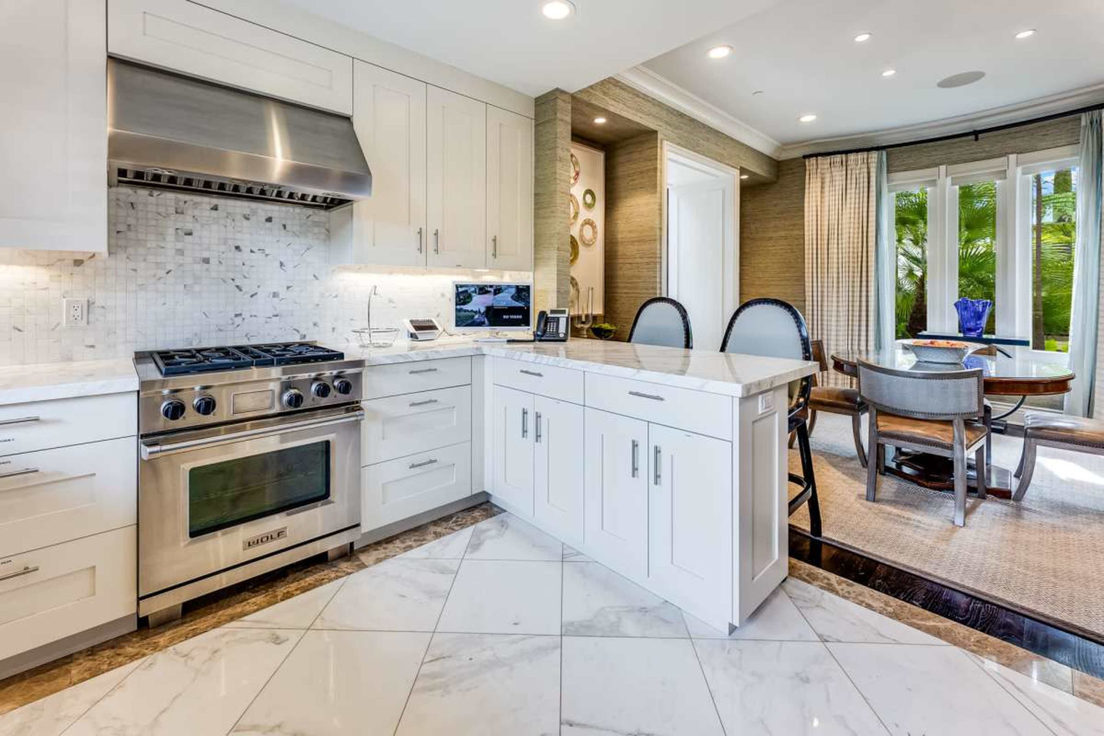 A modern kitchen features stainless steel appliances, white cabinetry, and a marble island, with a dining area visible through large windows.