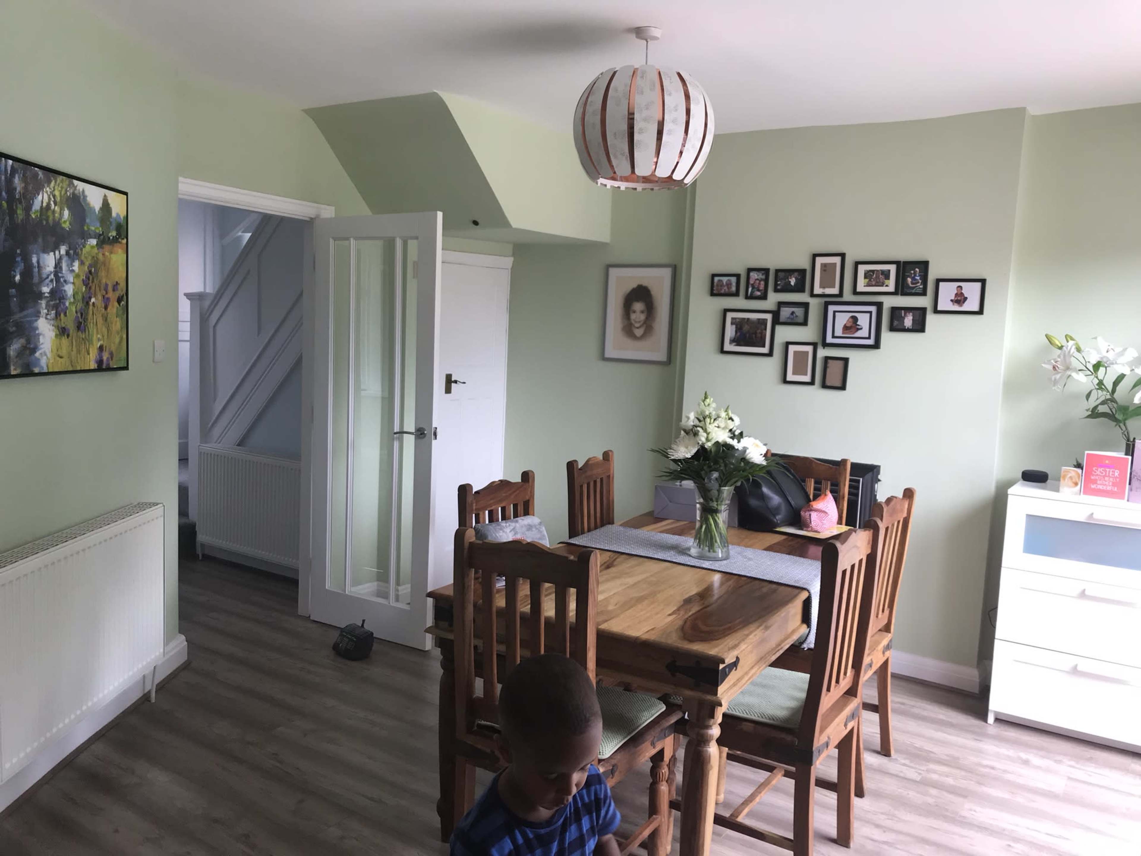 A dining area features a wooden table surrounded by chairs, a flower vase, and walls adorned with pictures, while an adjacent room can be seen through an open door.