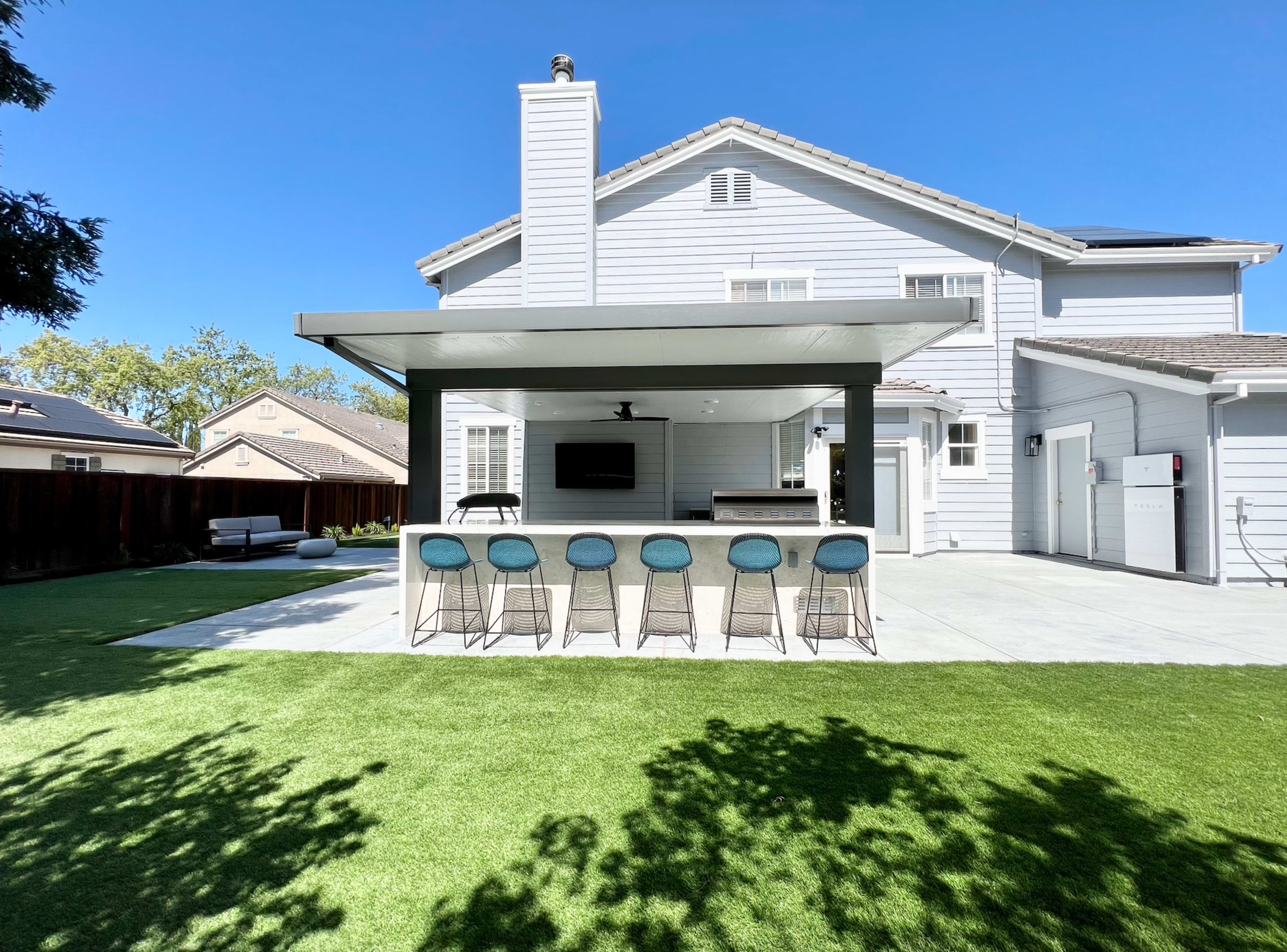 A modern backyard features a covered patio with a bar area and stools, set against a well-maintained lawn and a two-story house.