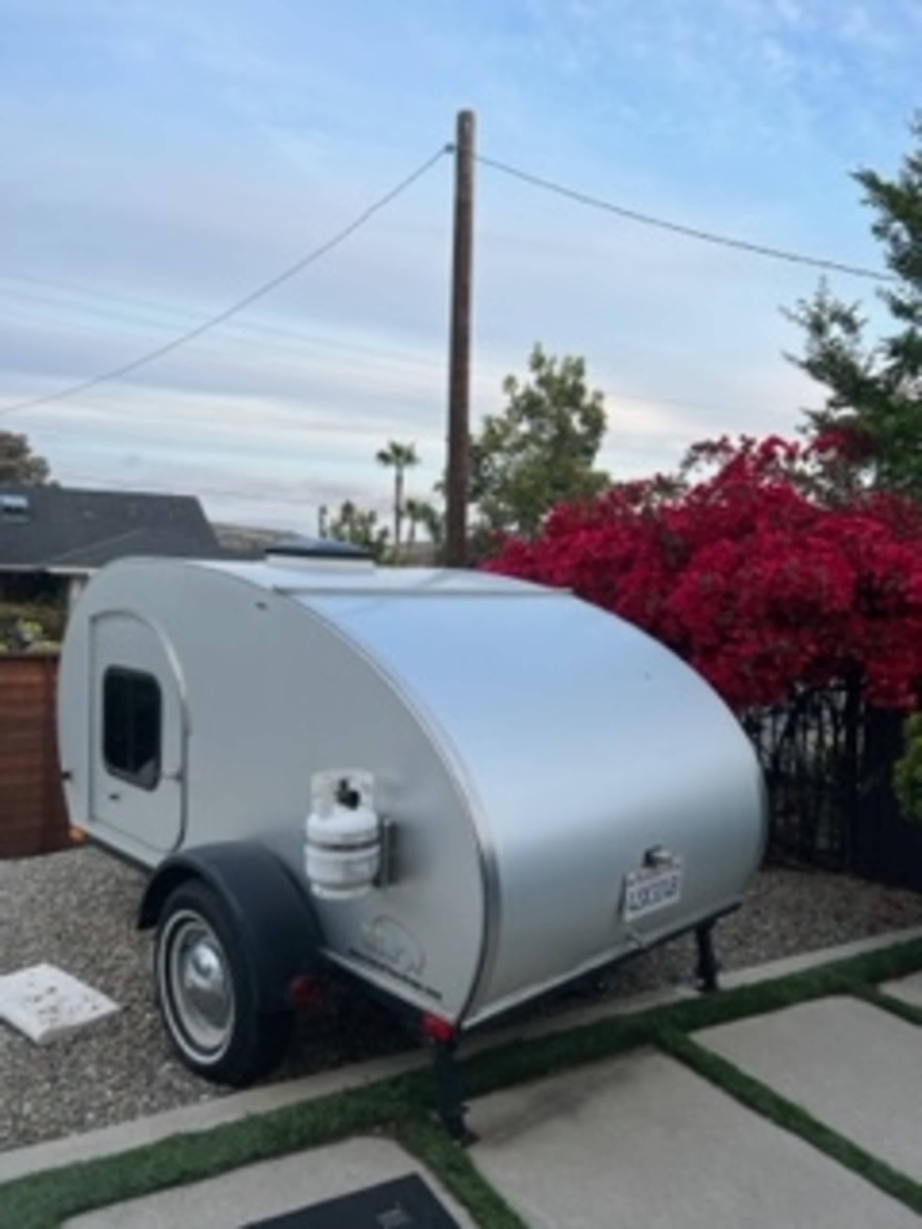 A silver teardrop trailer is parked beside a flowering bush and a concrete pathway.