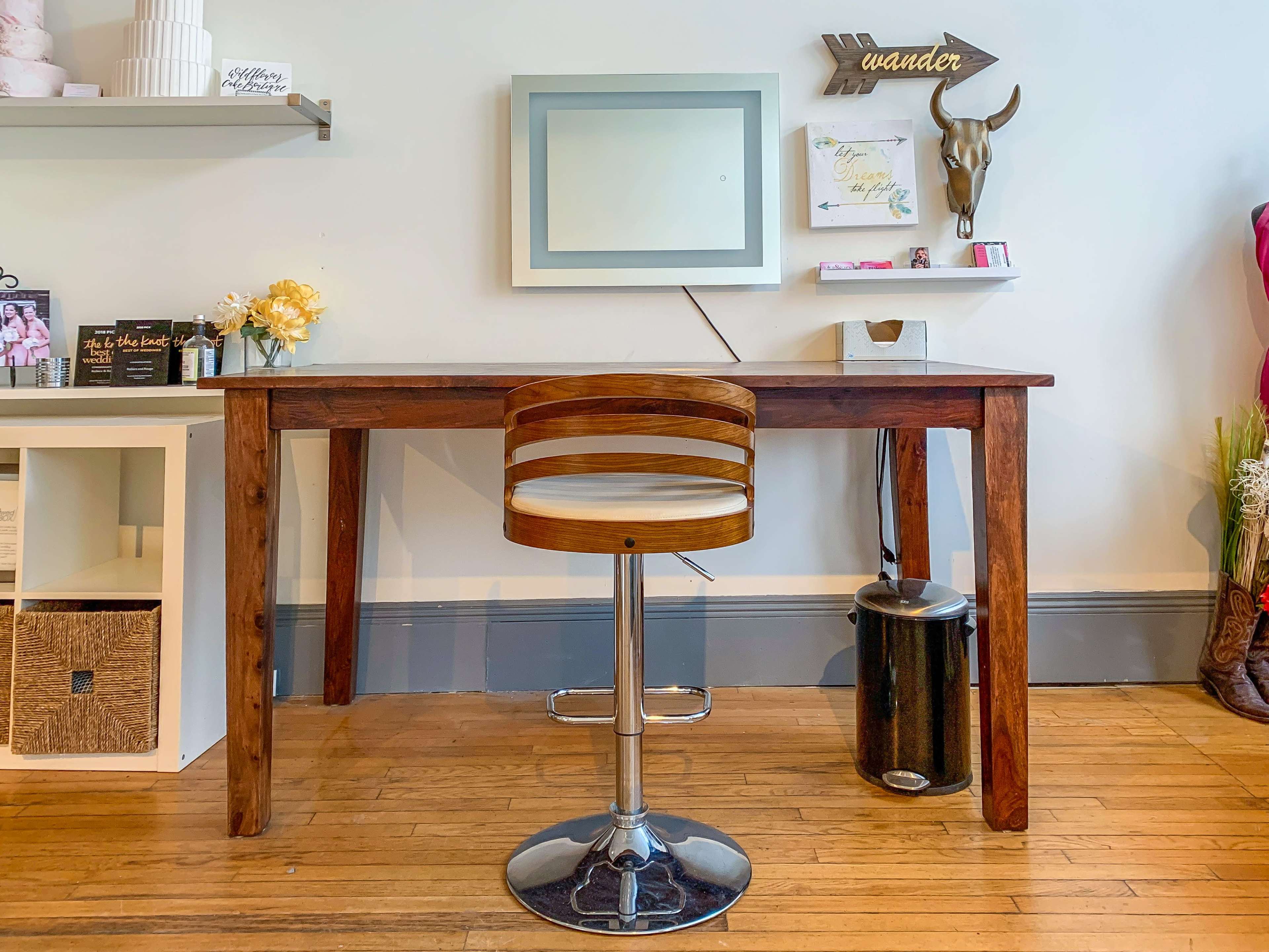 A wooden desk with a metal stool in front of a mirror, accompanied by decorative items and a trash bin in a well-lit room.