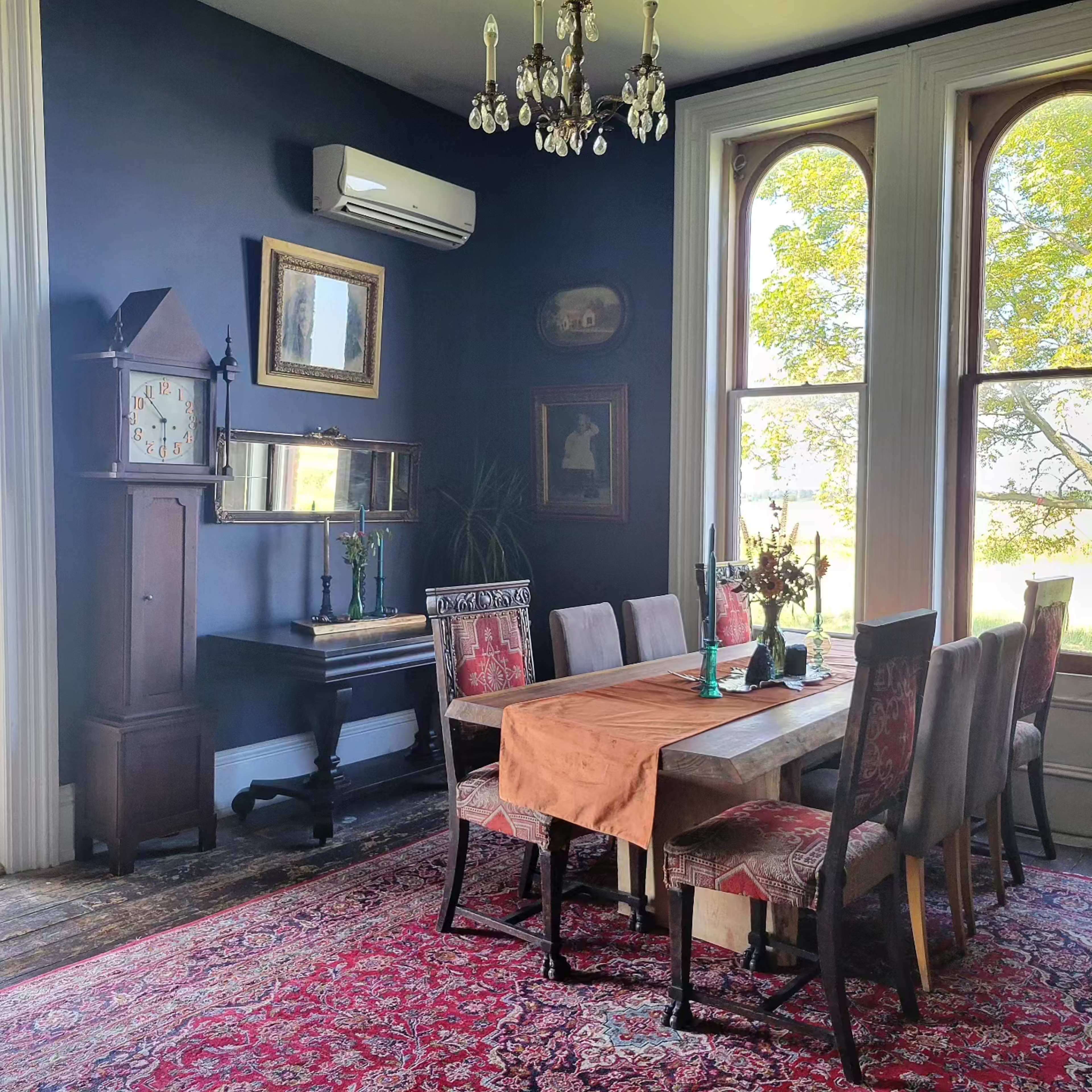 A dining room features a wooden table surrounded by upholstered chairs, with large windows casting light on vintage decor and a clock.
