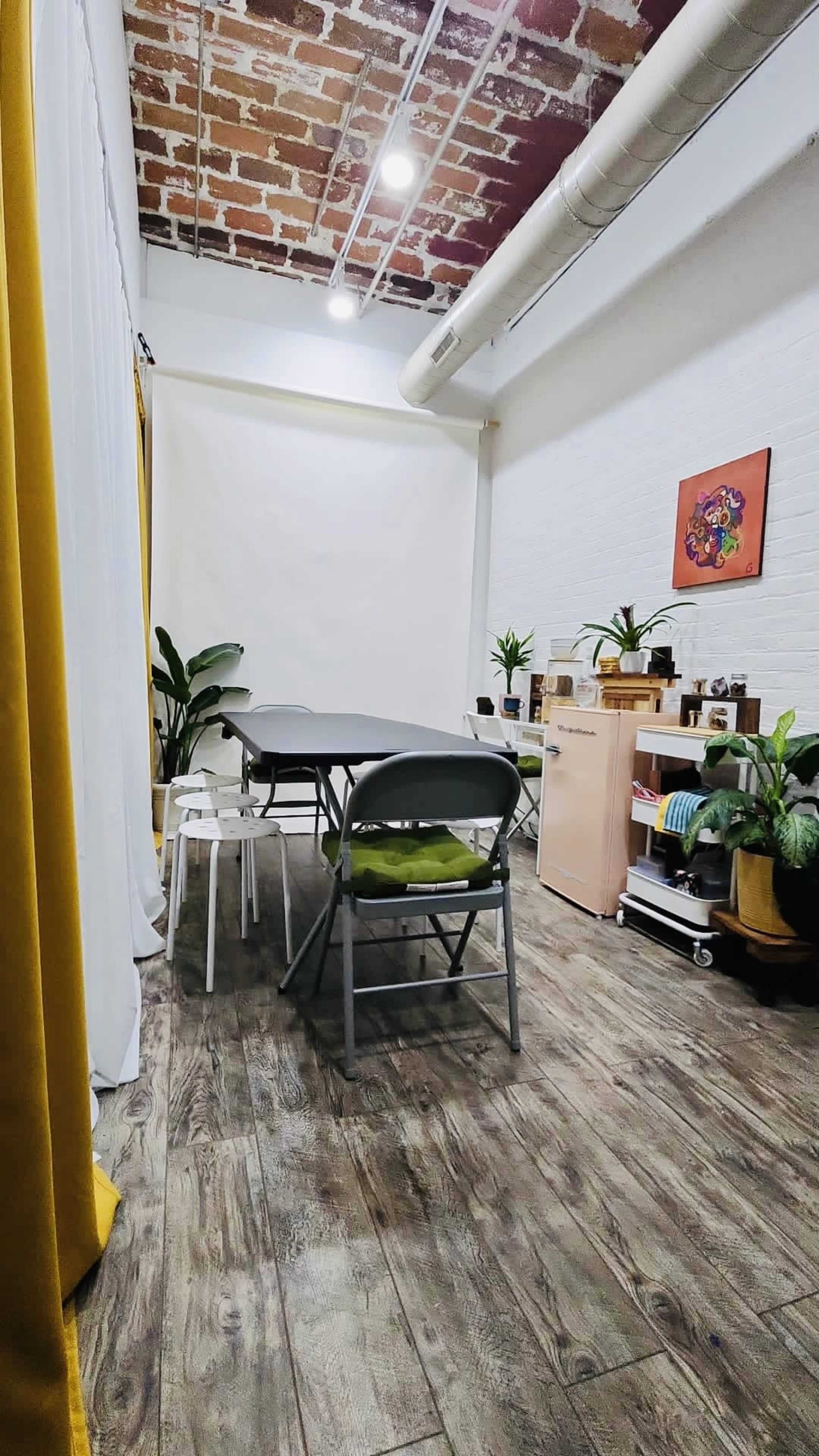 A bright, organized room with a long table, folding chairs, plants, and shelves lined with various items.