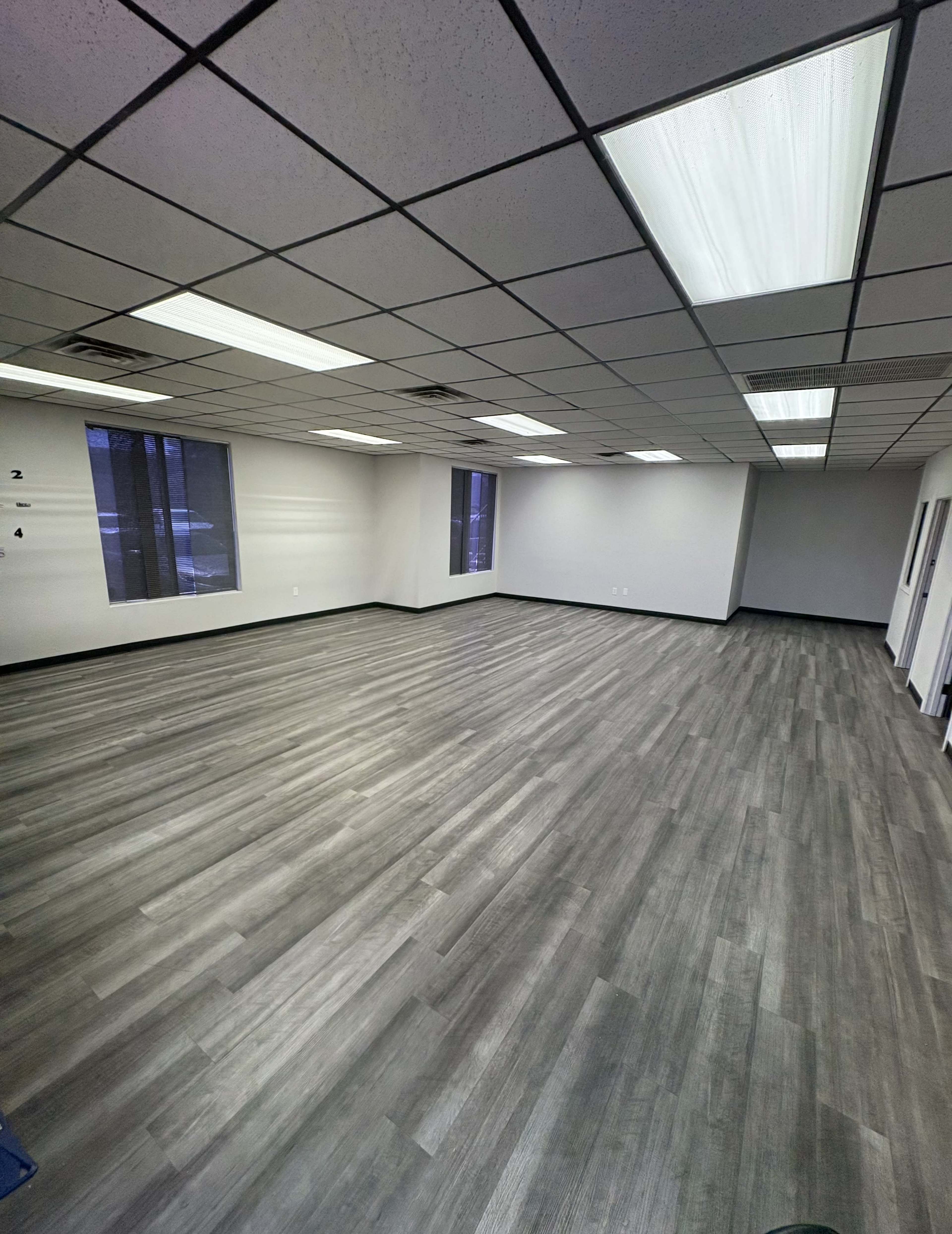 The image shows a large, empty room with gray laminate flooring and overhead fluorescent lighting.
