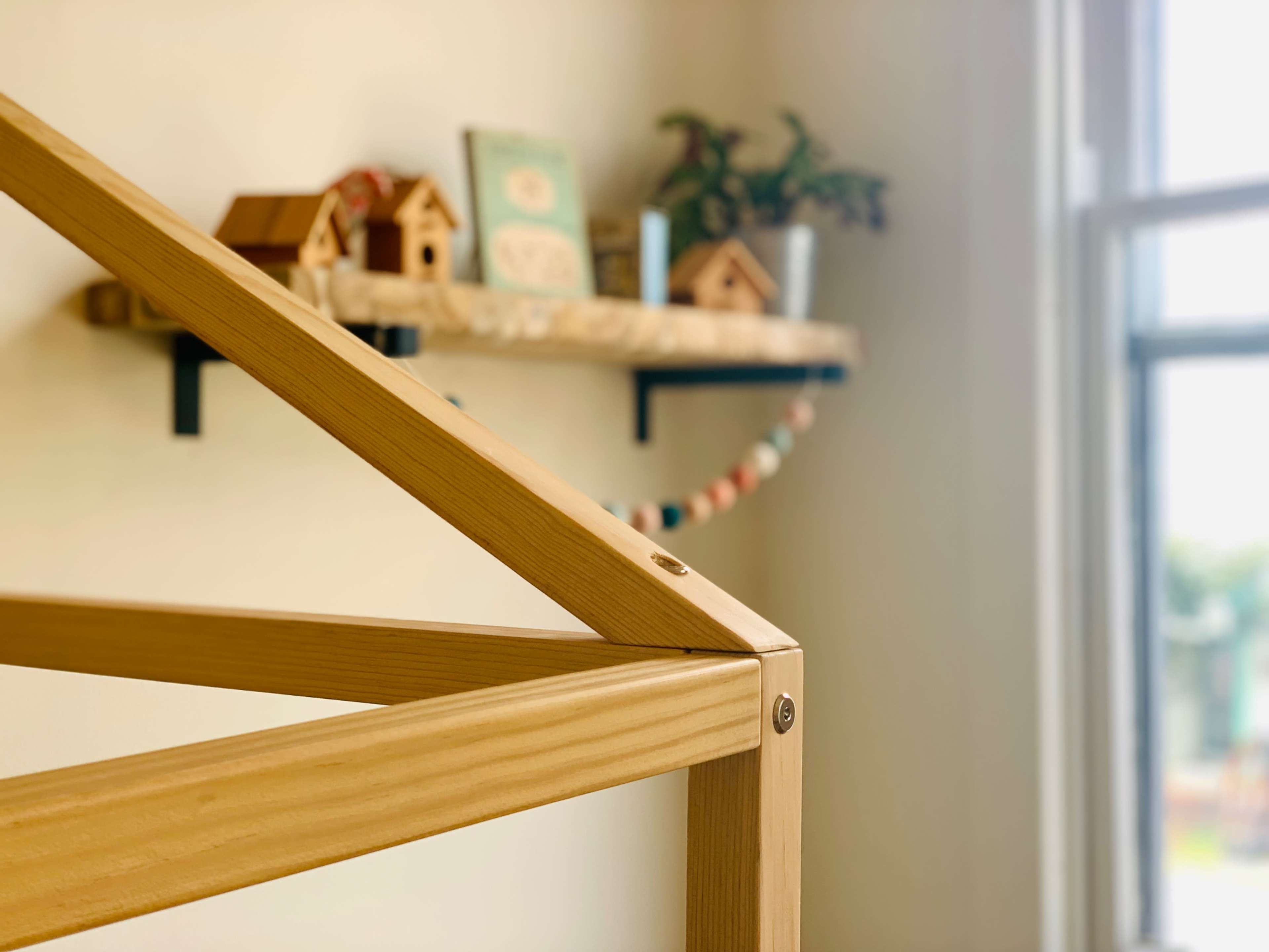 A wooden bed frame with an angled roof structure in the foreground, while a shelf with decorative items is visible in the background.