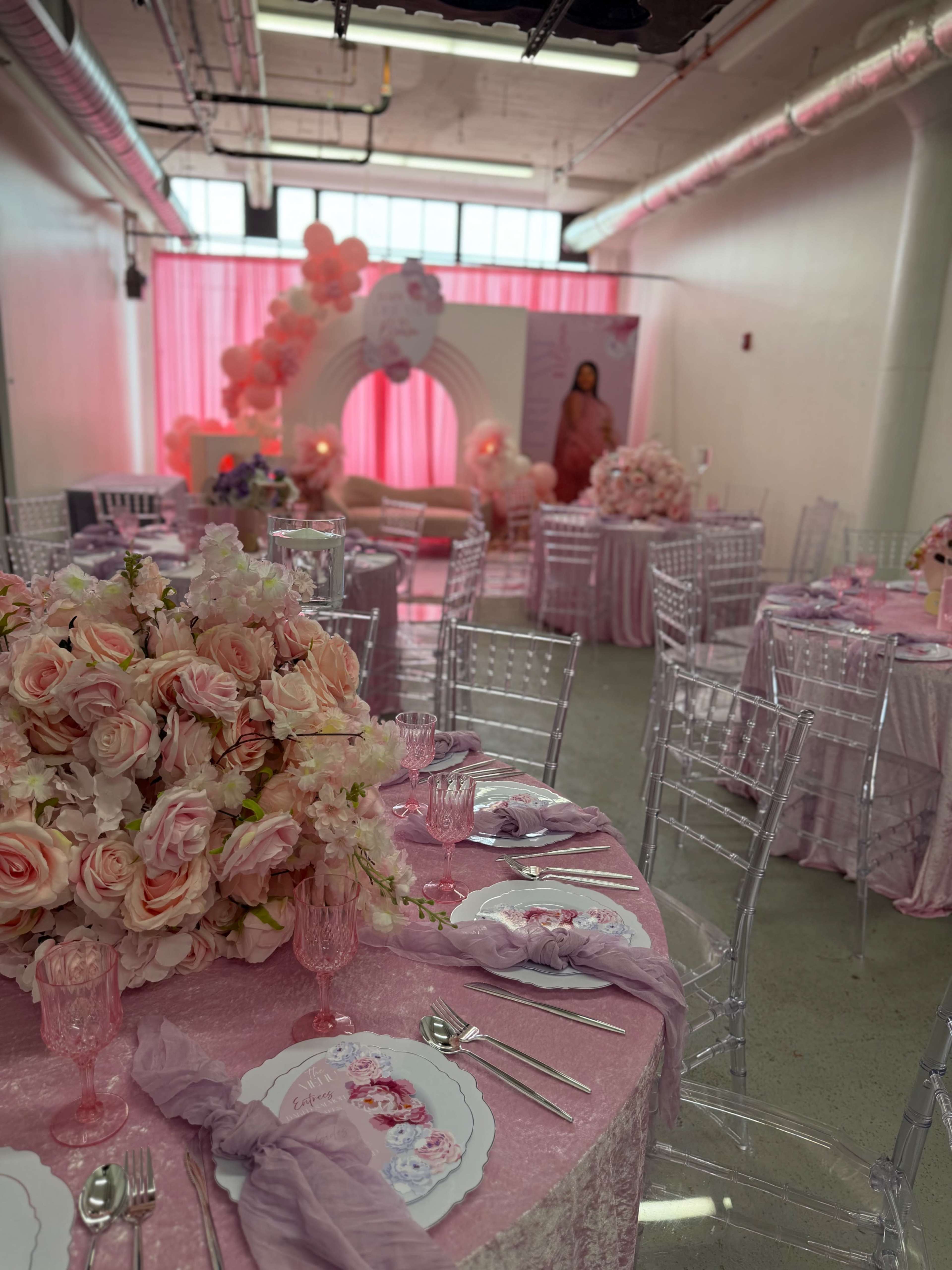 The image shows a banquet hall decorated for an event, featuring tables adorned with pink tablecloths, floral centerpieces, and a backdrop of pink drapes and balloons.
