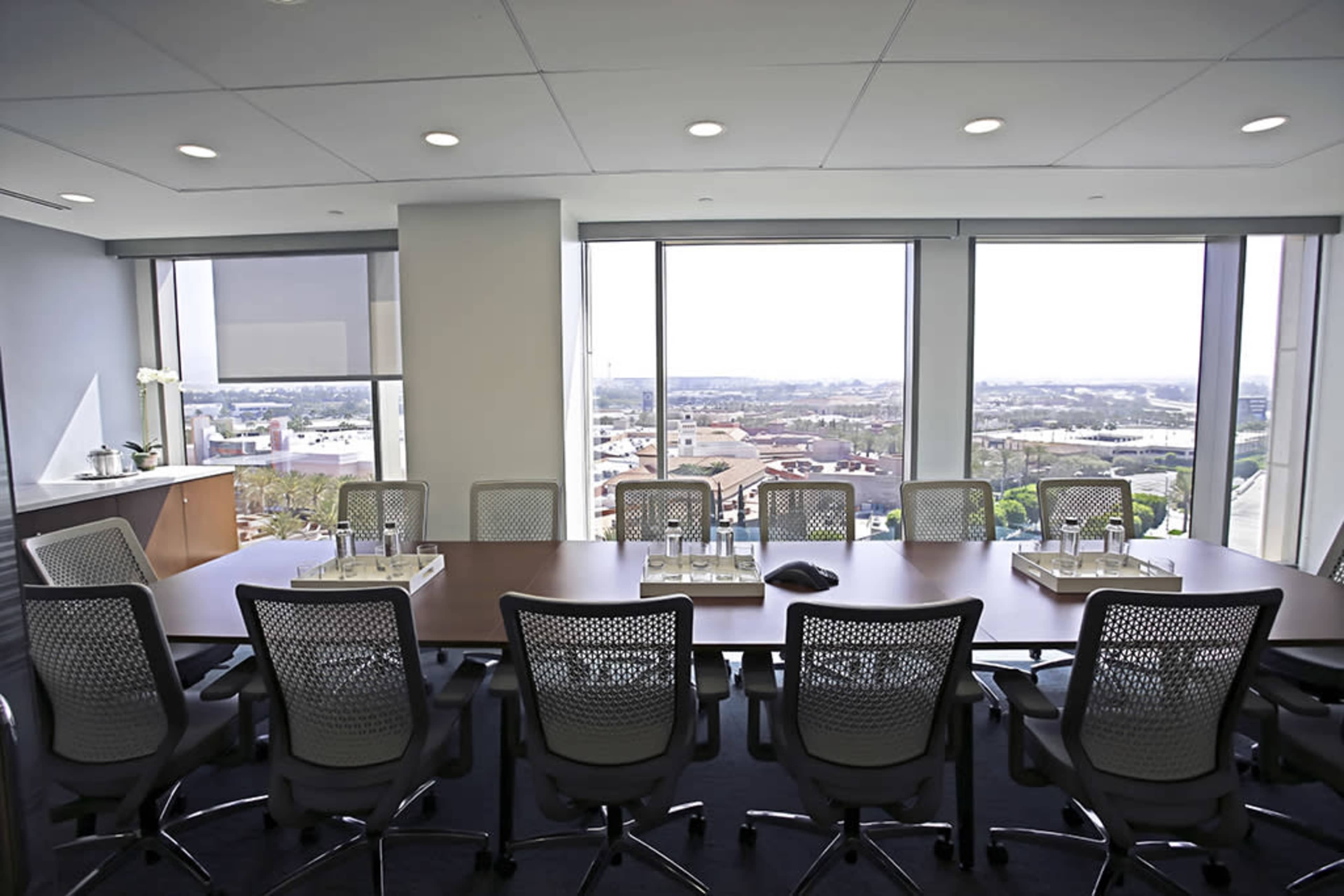 A modern conference room with a large wooden table surrounded by ergonomic chairs and large windows overlooking an urban landscape.