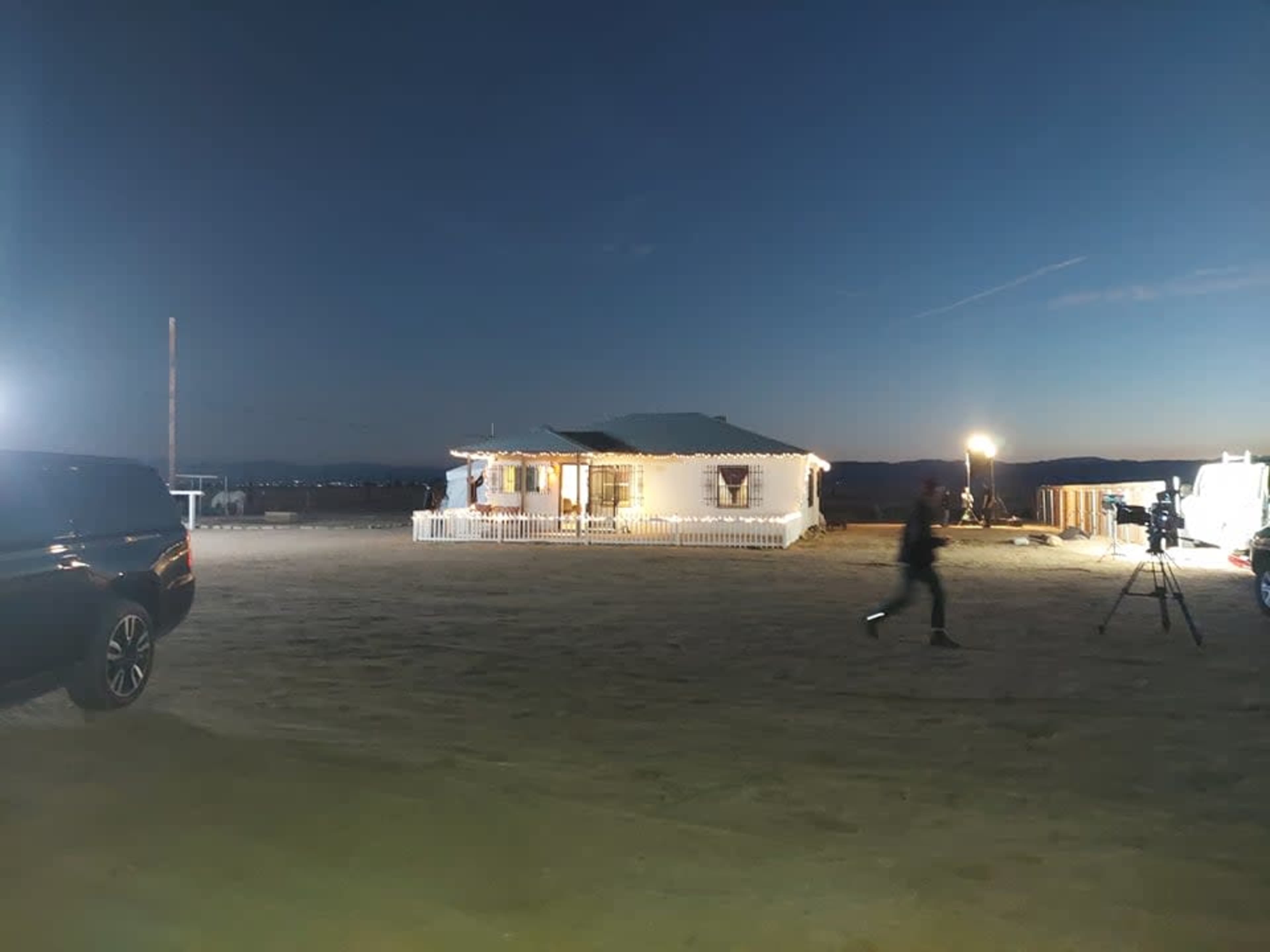 A brightly lit house stands in a sandy outdoor area during twilight, with a person walking past and filming equipment nearby.
