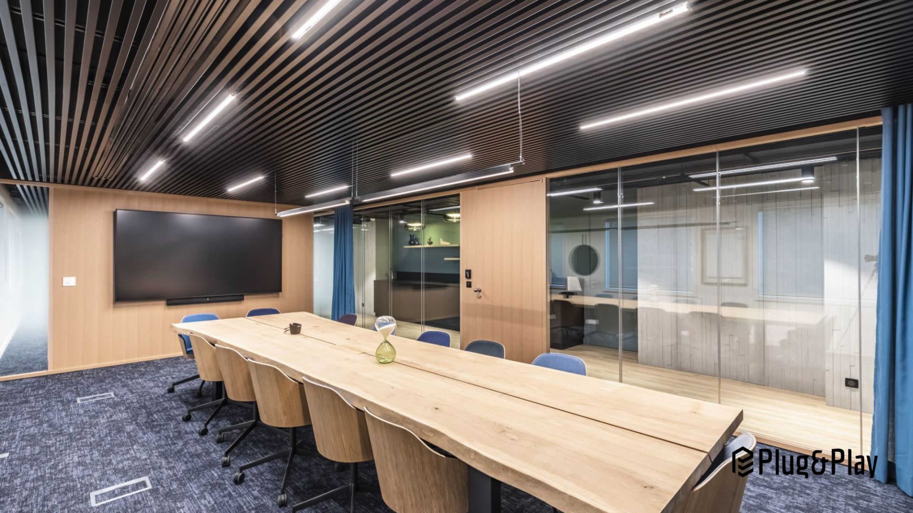 The image shows a modern conference room with a long wooden table, ergonomic chairs, a large screen on the wall, and glass walls providing visibility into adjacent spaces.