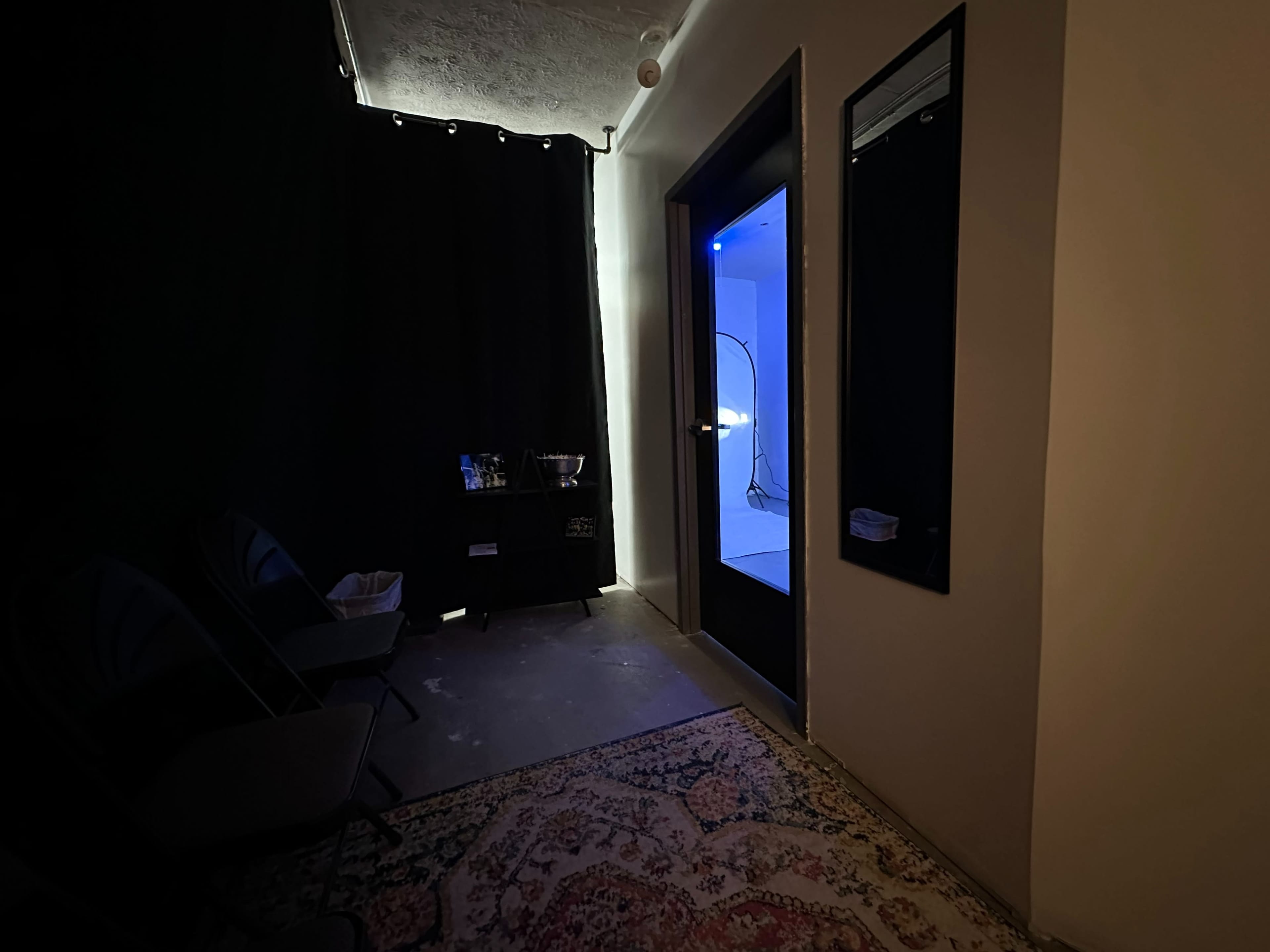 A dimly lit hallway with a doorway leading to a blue-lit room, flanked by a black curtain and a small furniture piece.