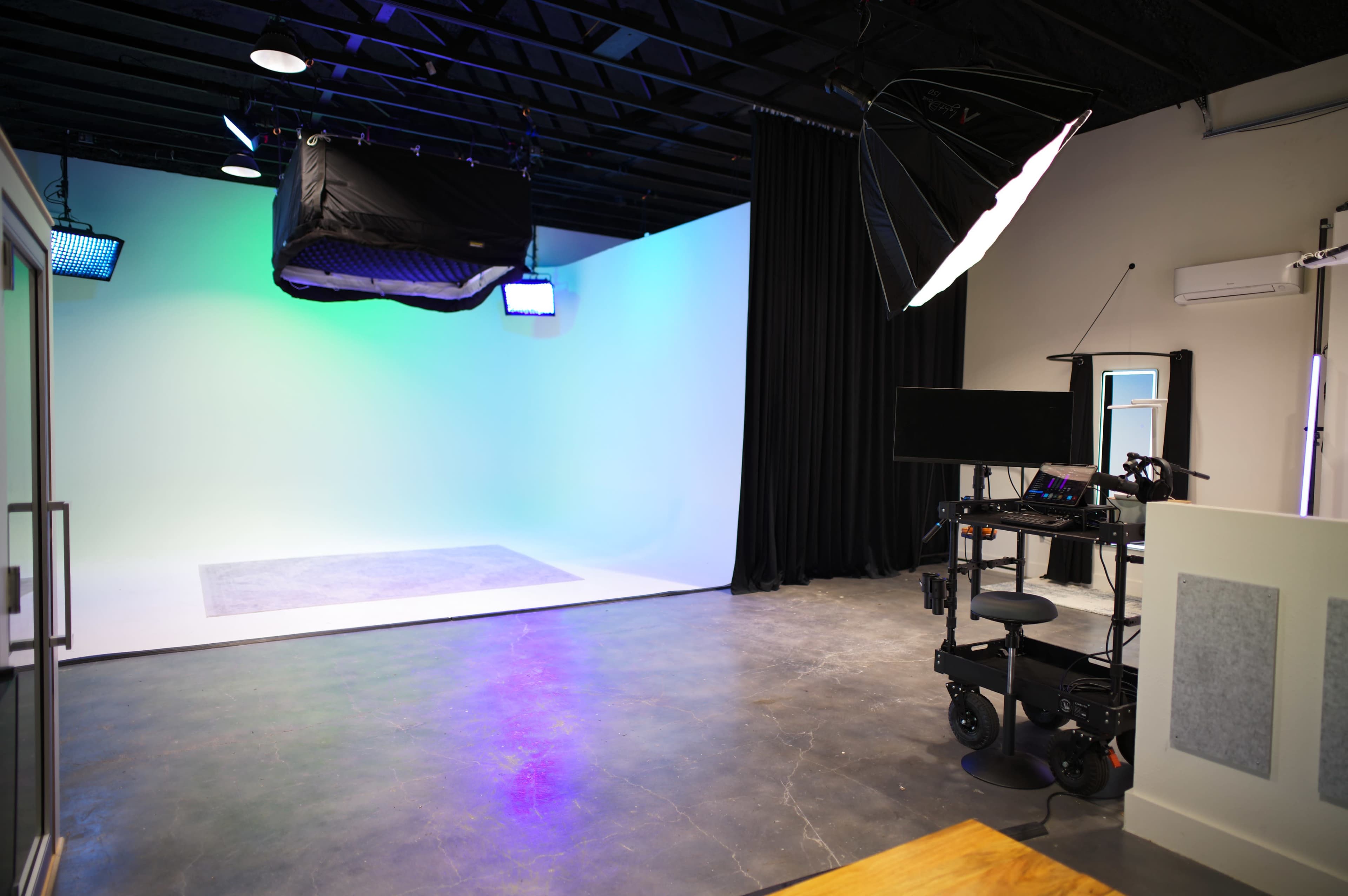 The image shows a professional photography or video studio with a seamless backdrop, lighting equipment, and a mobile workstation.