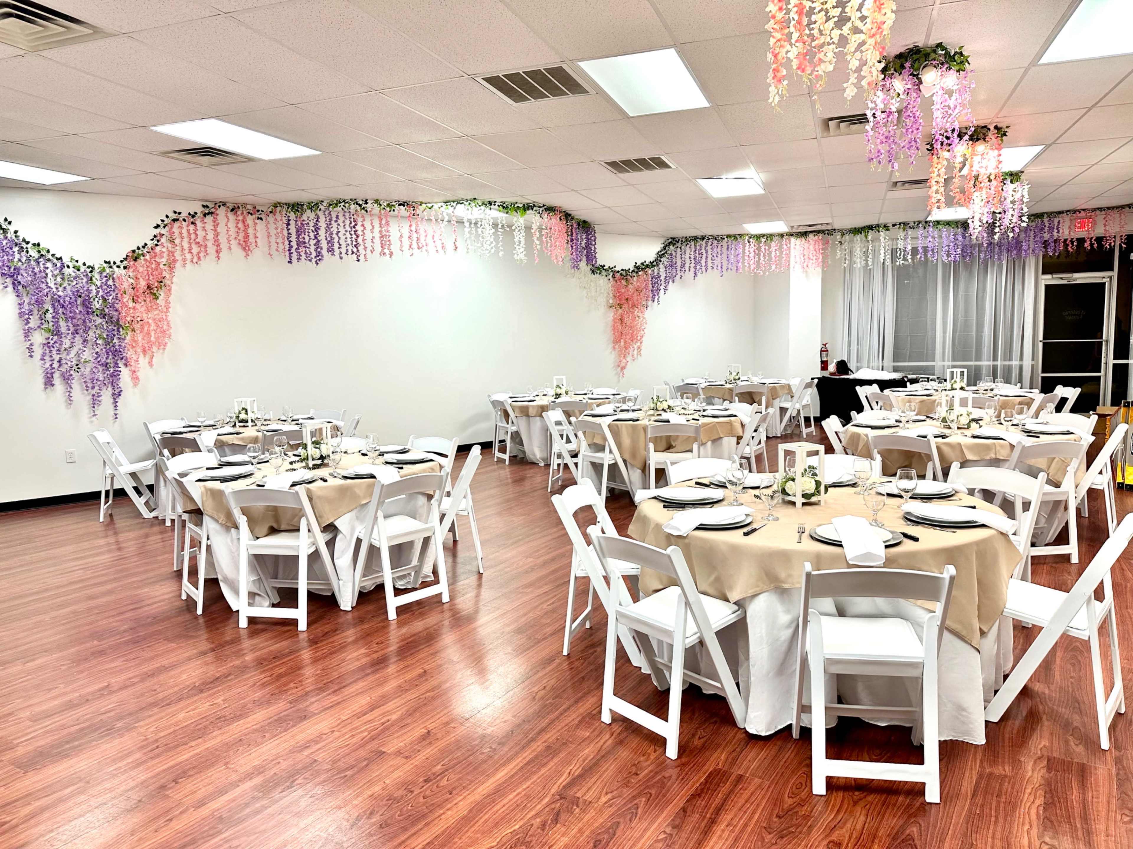 A banquet hall features several round tables covered with beige tablecloths, surrounded by white folding chairs, and adorned with colorful decorations hanging from the ceiling.