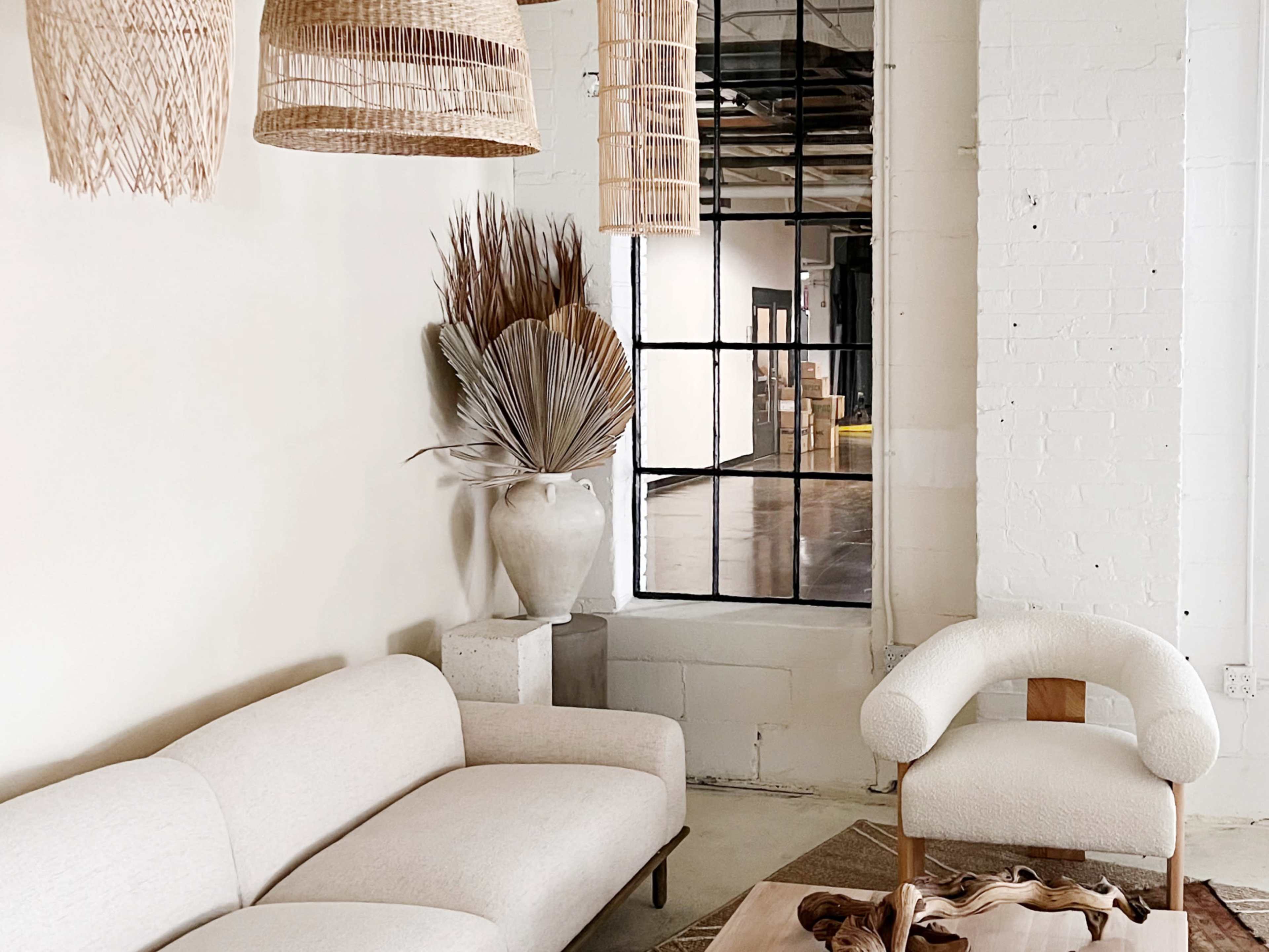 The image displays a minimalist interior featuring a light-colored sofa, a textured armchair, woven pendant lights, and a decorative arrangement of dried plants in a ceramic vase.