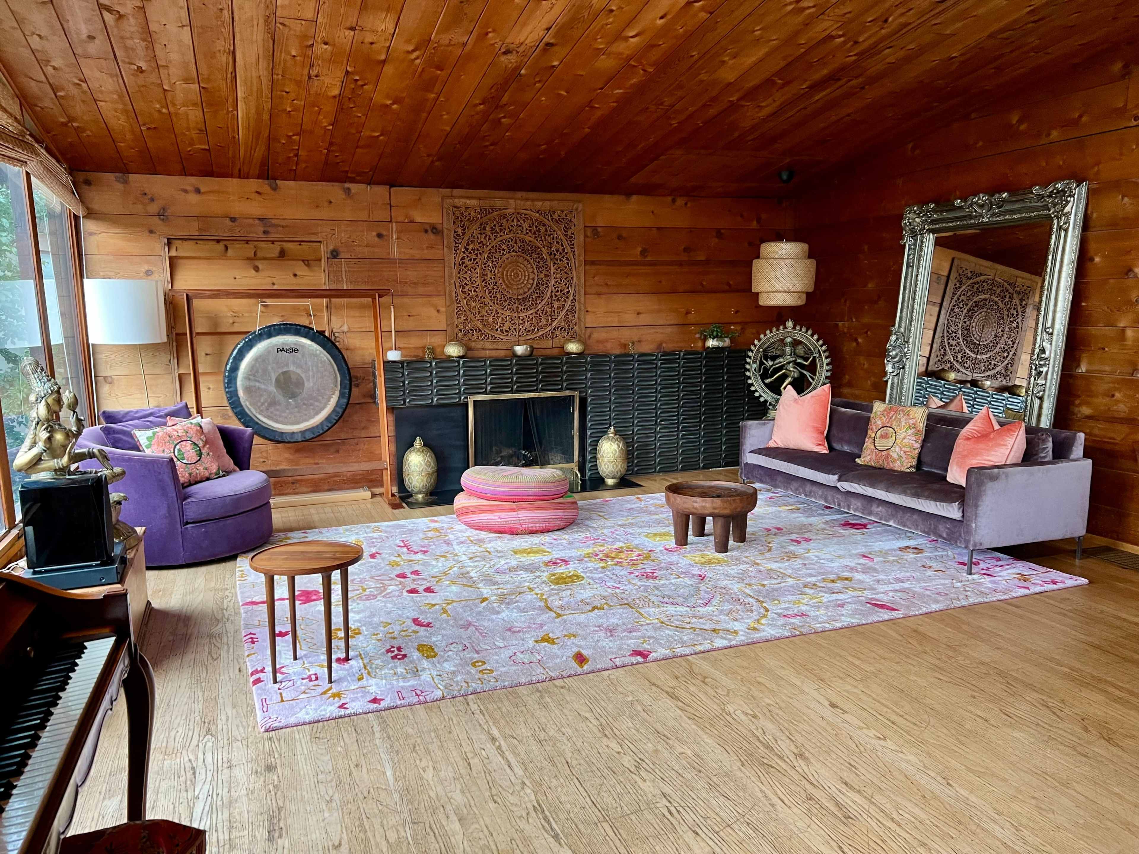 A well-decorated living room with wooden walls, a large pink area rug, a fireplace, and various decorative elements including a gong and a large mirror.