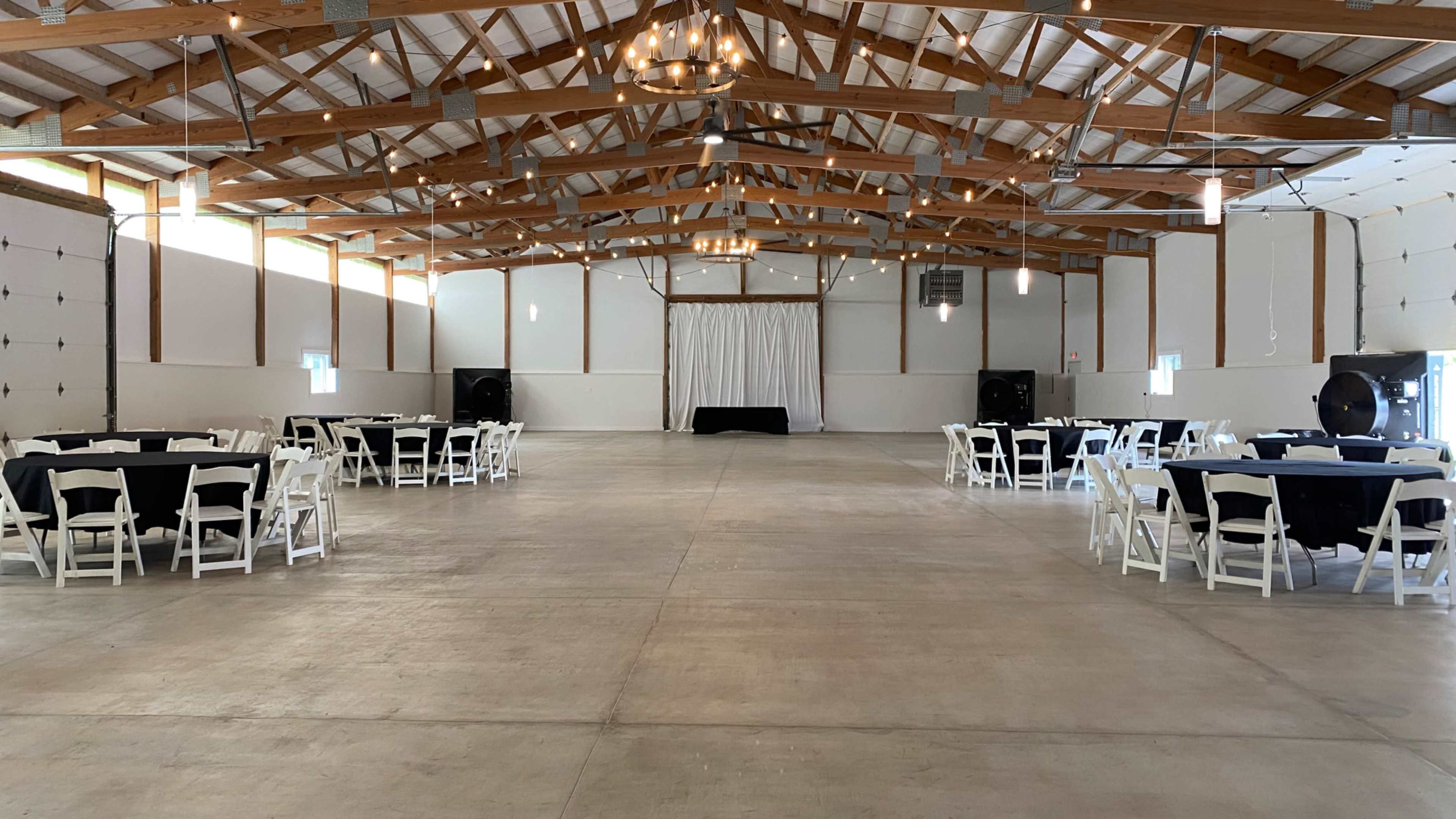 The image shows a spacious hall with wooden beams, featuring several round tables set with white tablecloths and black accents, and a stage area at the front.