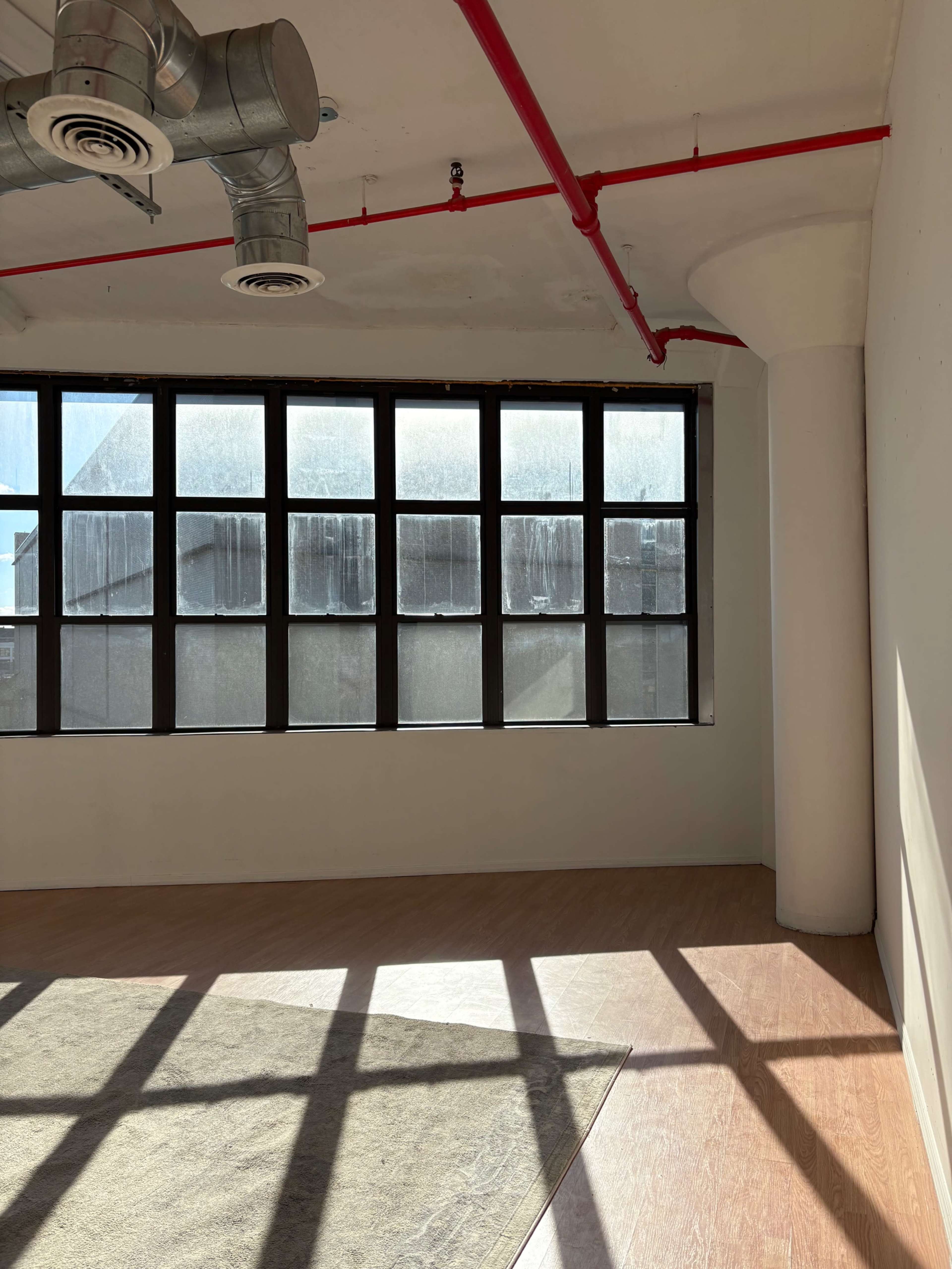 A modern, empty room features large windows with industrial-style ducts above and sunlight casting shadows on the wooden floor.