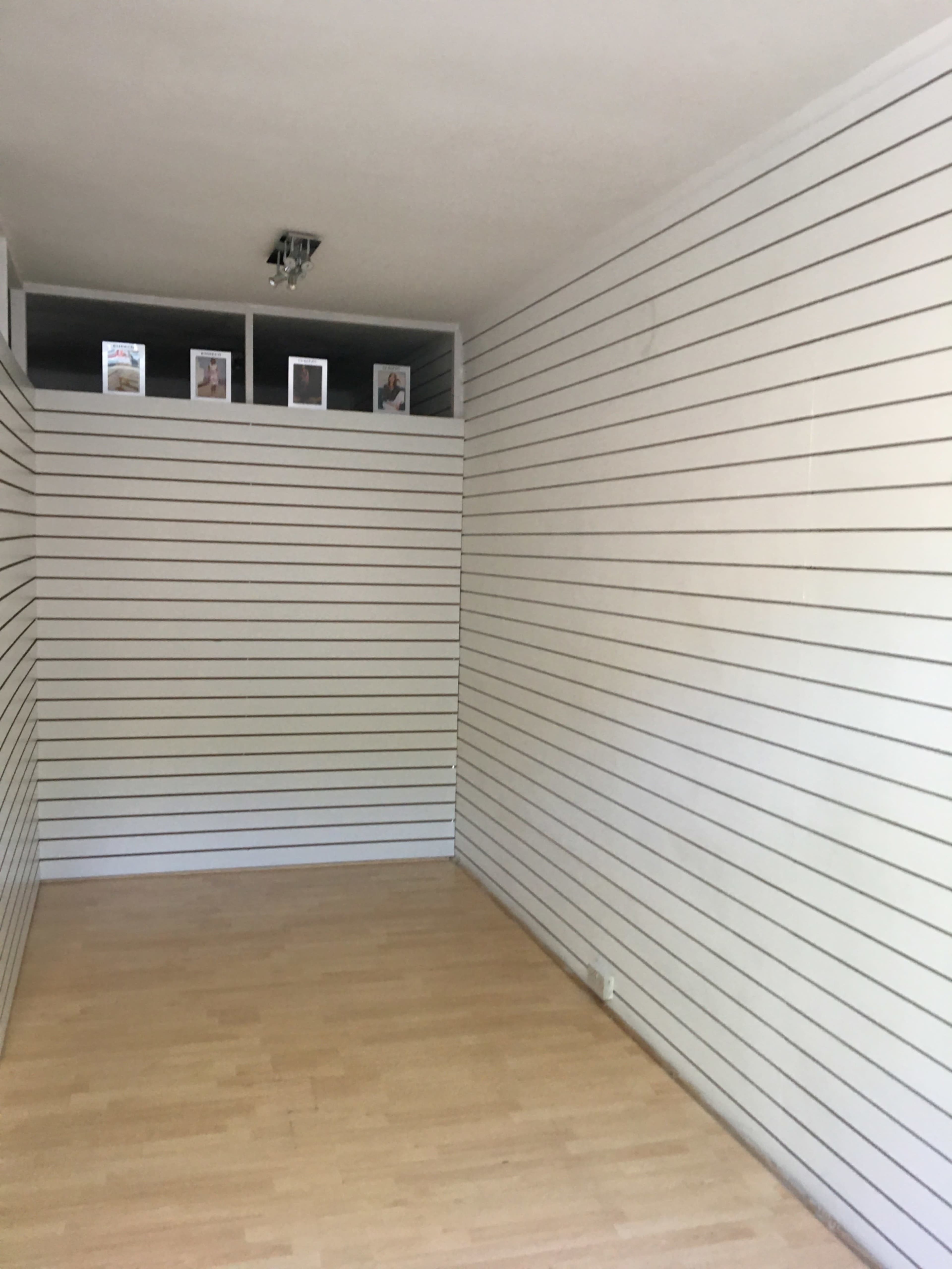 The image shows an empty room with wooden flooring and white striped walls, featuring a shelf at the back displaying a few framed pictures.