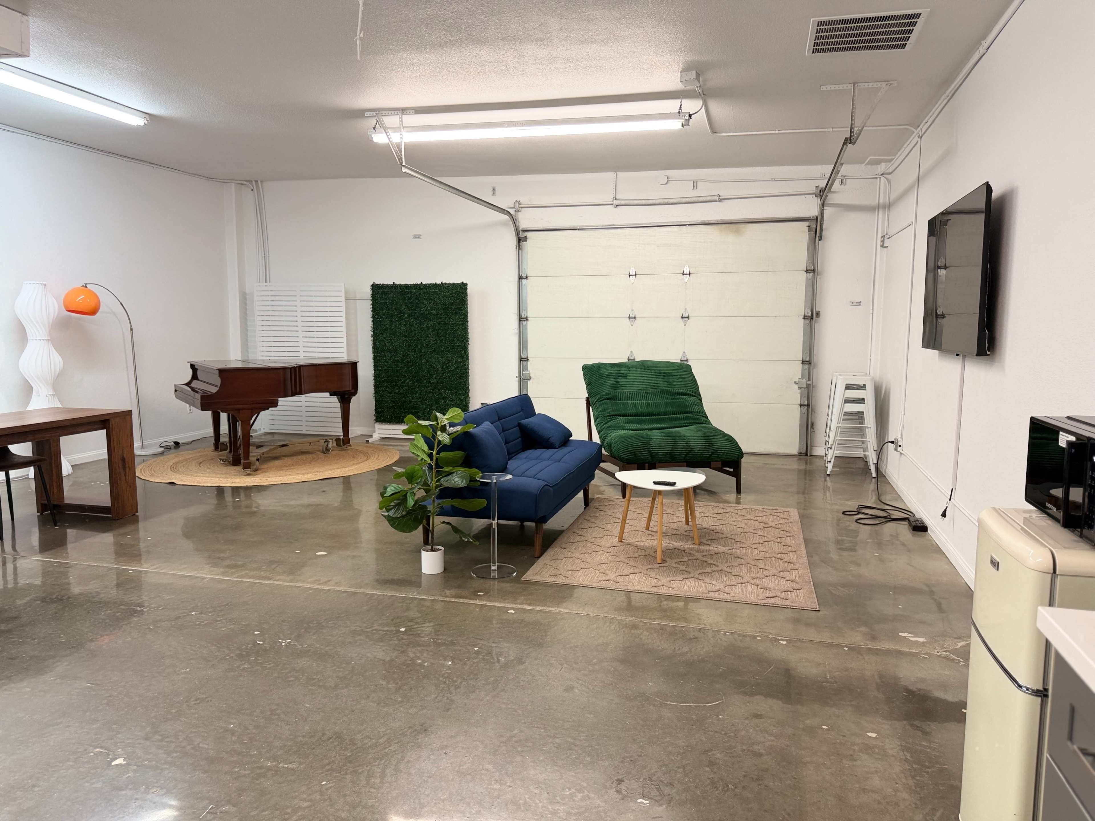 The image shows a spacious, minimalist room with polished concrete floors, featuring a blue sofa, a green lounge chair, a piano, a small table, and a plant, along with garage doors at one end.