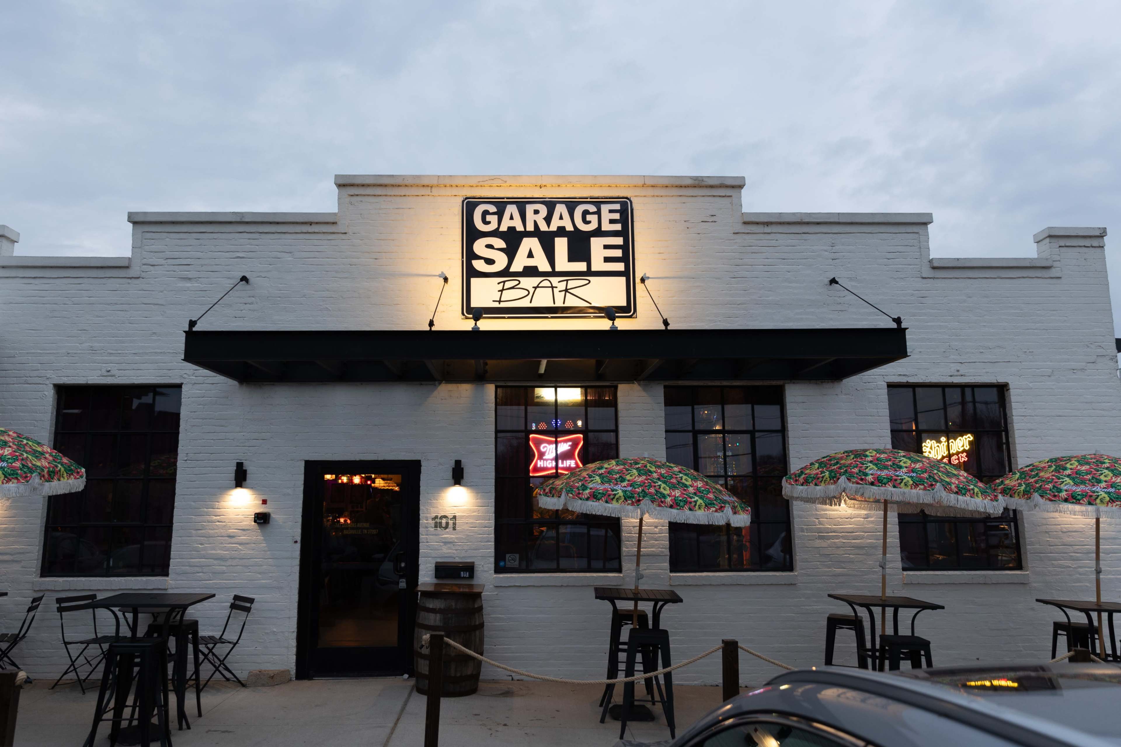 The image shows the exterior of a bar labeled "Garage Sale Bar" with outdoor seating and colorful umbrellas.