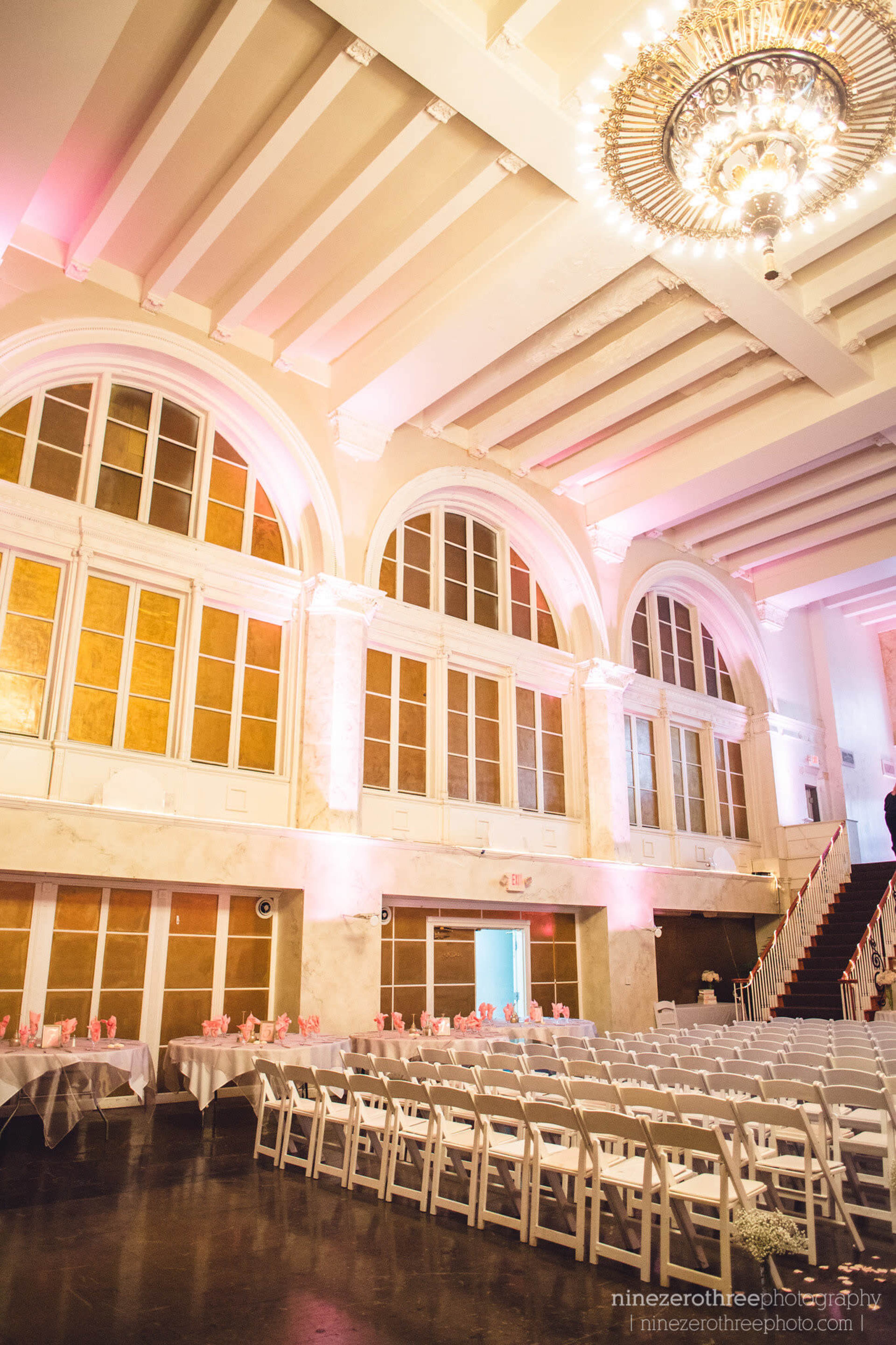 The image shows a spacious, elegantly decorated indoor venue with arched windows, a chandelier, and rows of tables set up for an event.