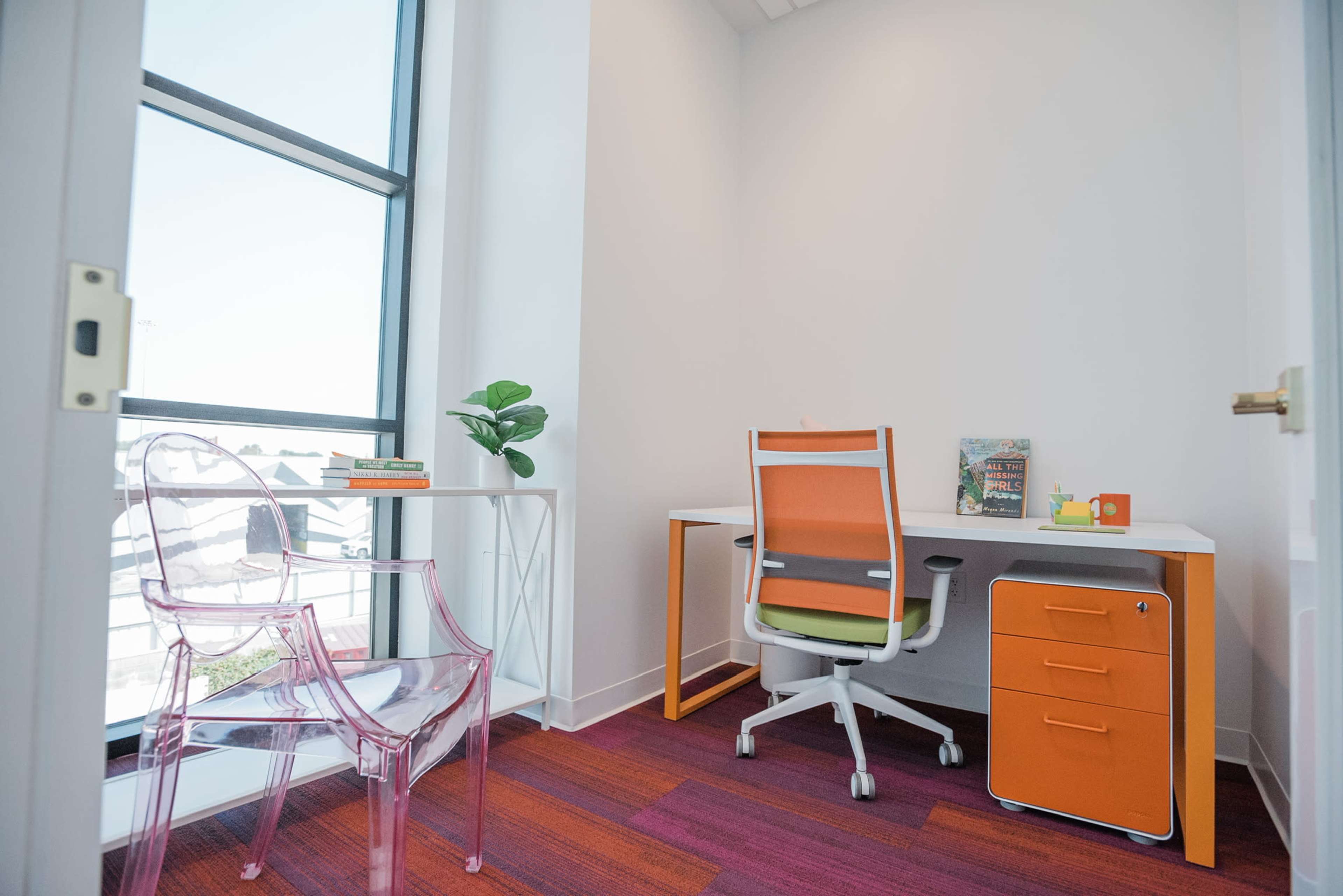 The image shows a small office space with a desk, a chair, a colorful accent chair, and a window providing natural light.