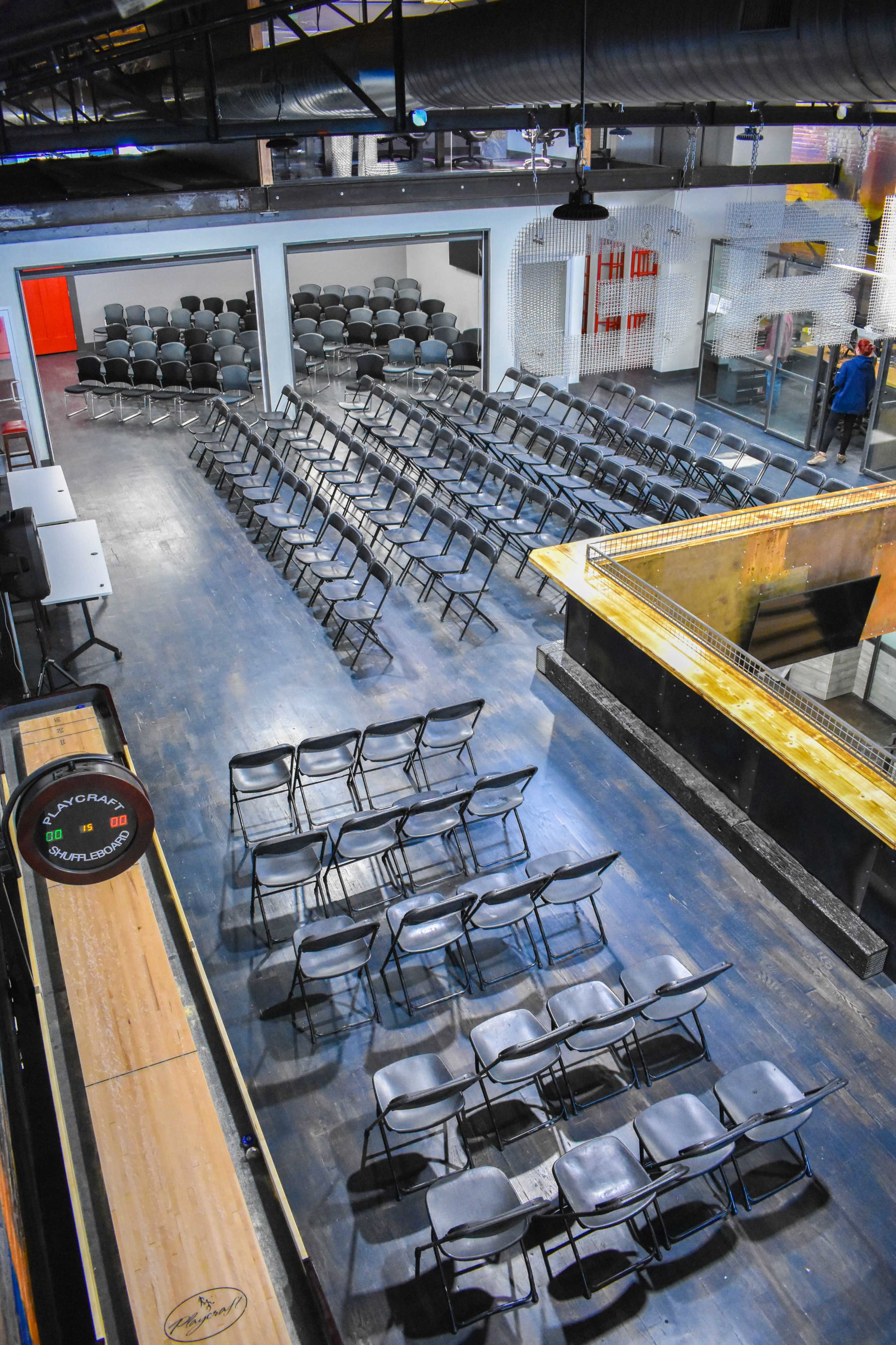 The image shows a spacious indoor venue set up with rows of black folding chairs arranged in front of a bar area, with large windows and an open layout.