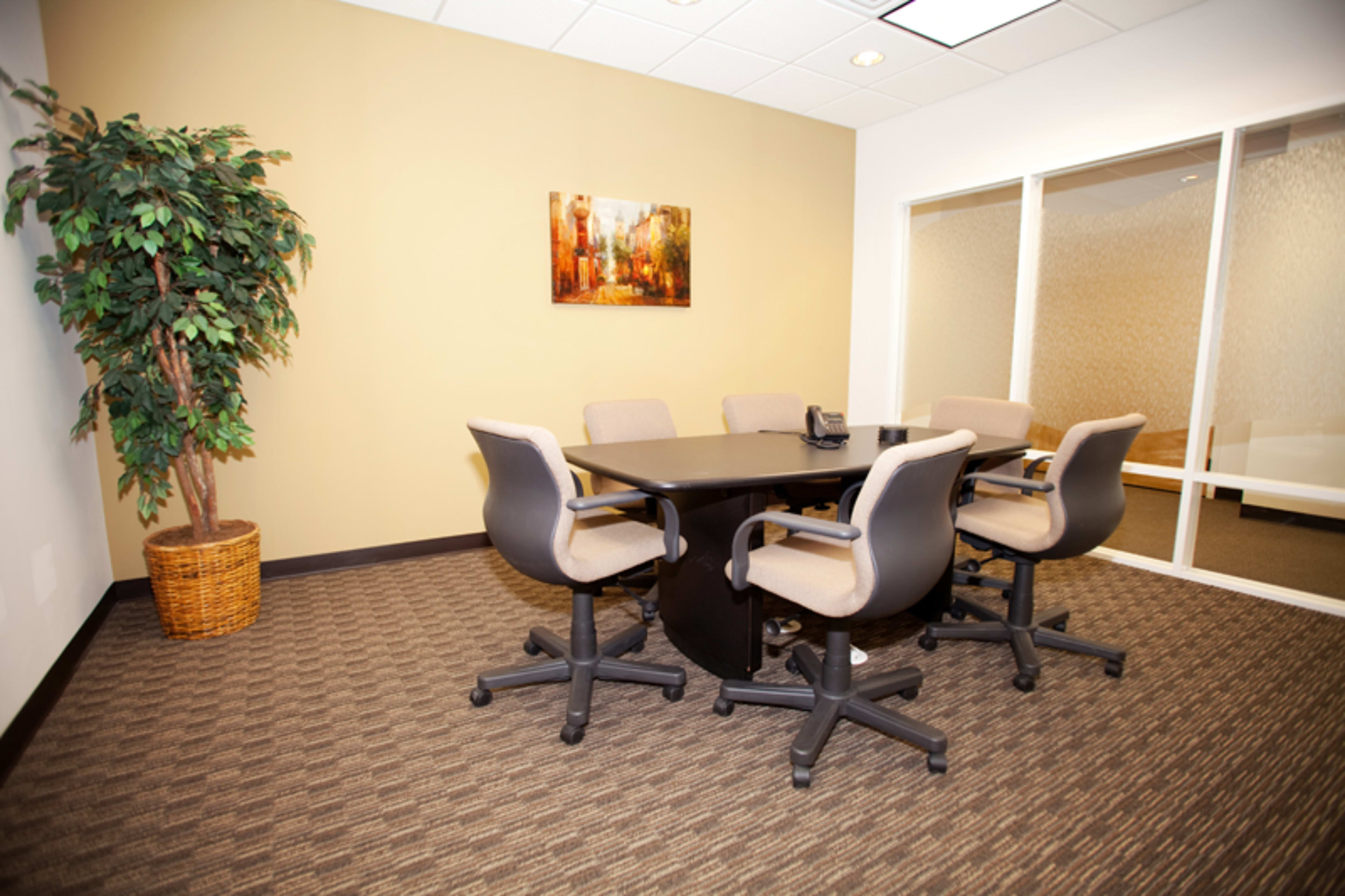 A conference room with a round black table surrounded by five wheeled office chairs, a potted plant in the corner, and a framed painting on the wall.
