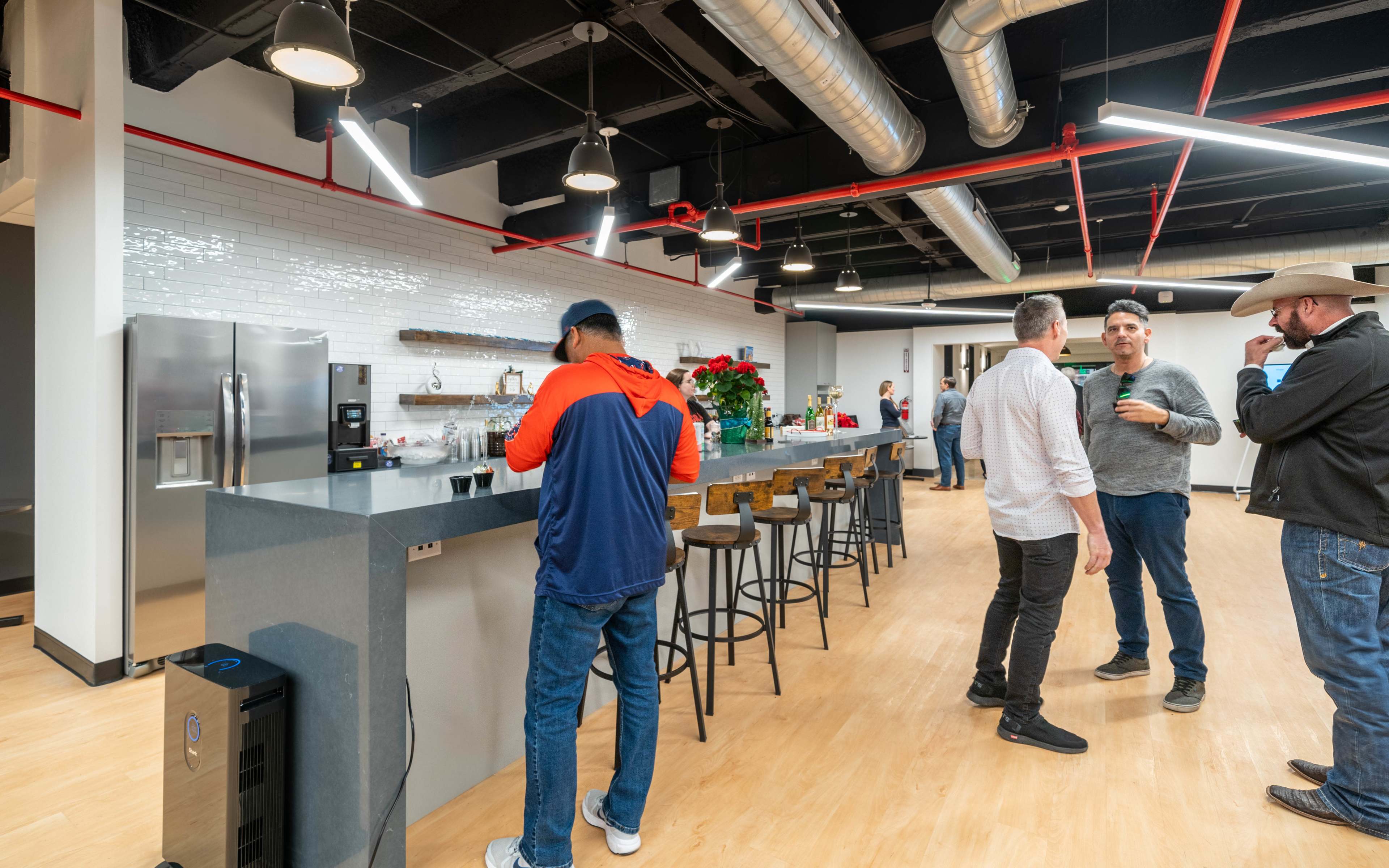 A modern kitchen space features a long countertop with bar stools, stainless steel appliances, and several people engaged in conversation.