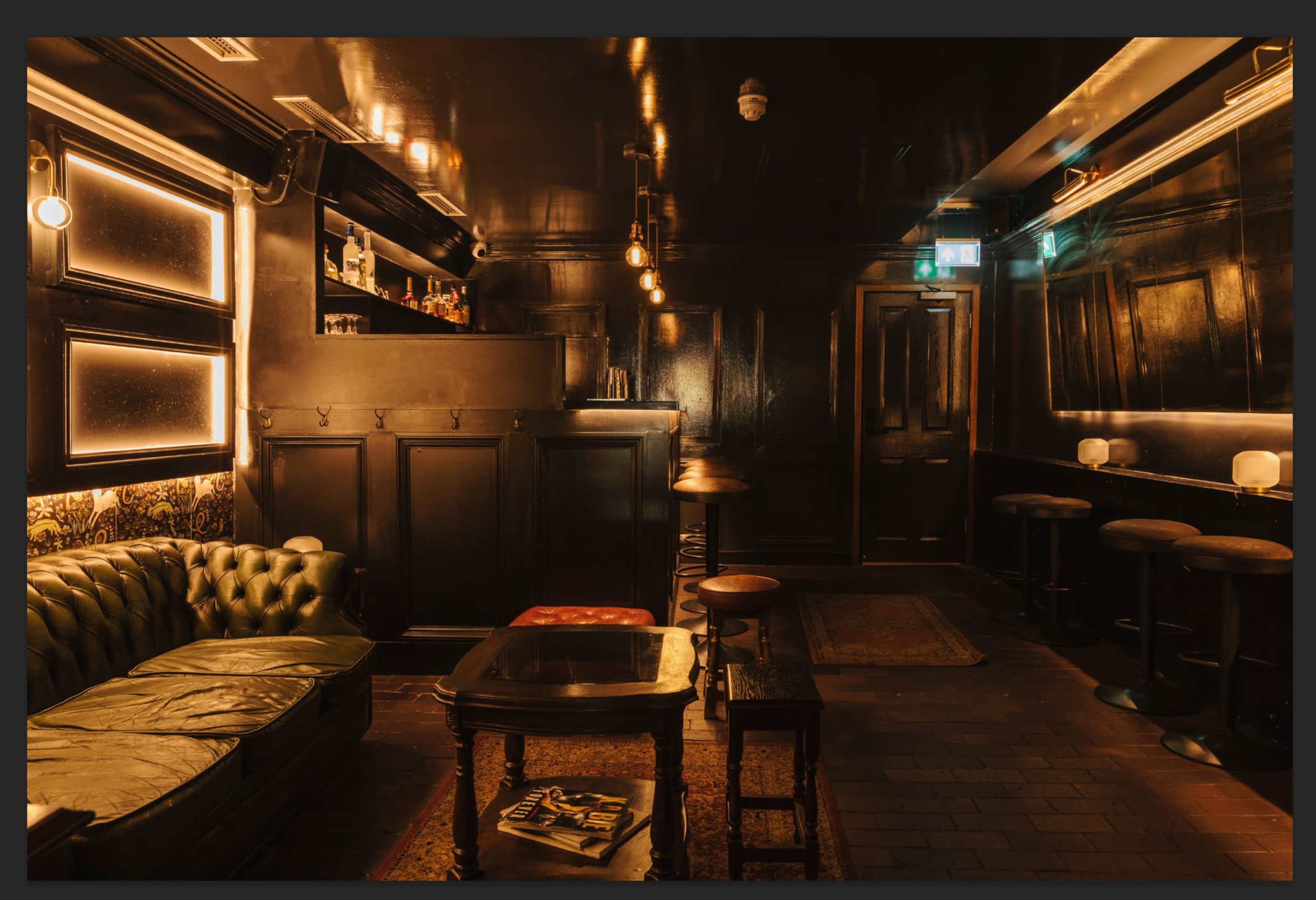 A dimly lit bar features a leather couch, wooden tables, and a polished counter with shelves of bottles.