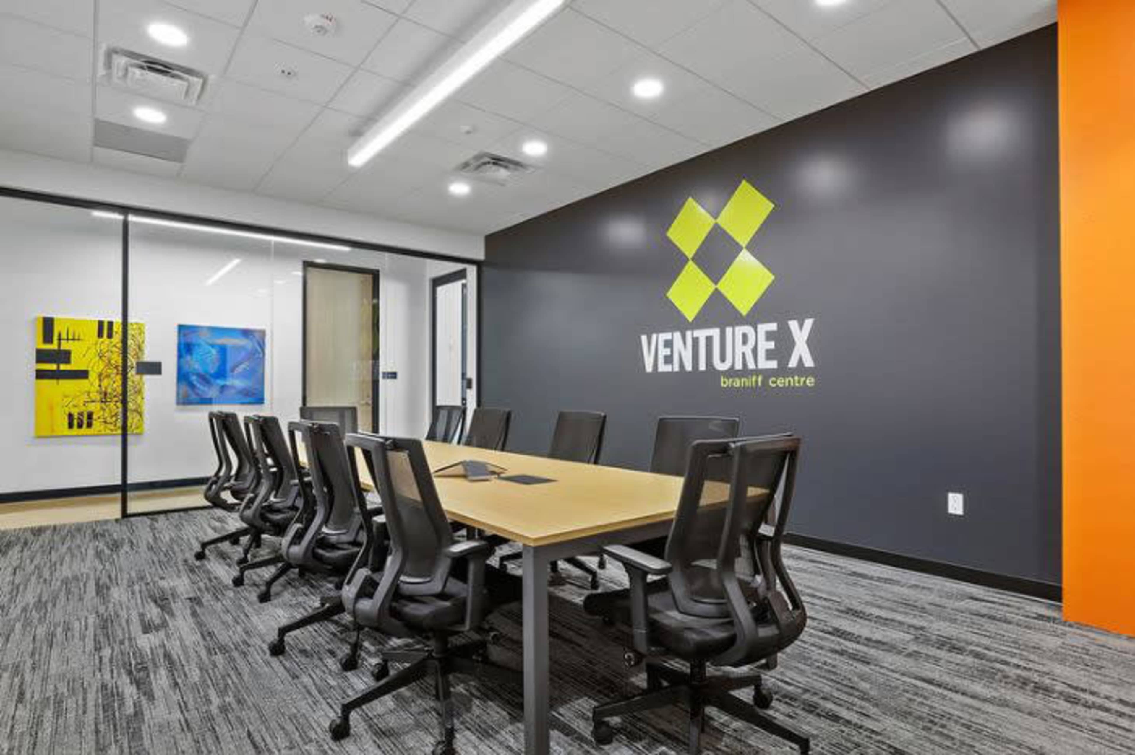 The image shows a modern conference room with a long table surrounded by ergonomic chairs, featuring a green and yellow logo on the wall and artwork displayed in frames.