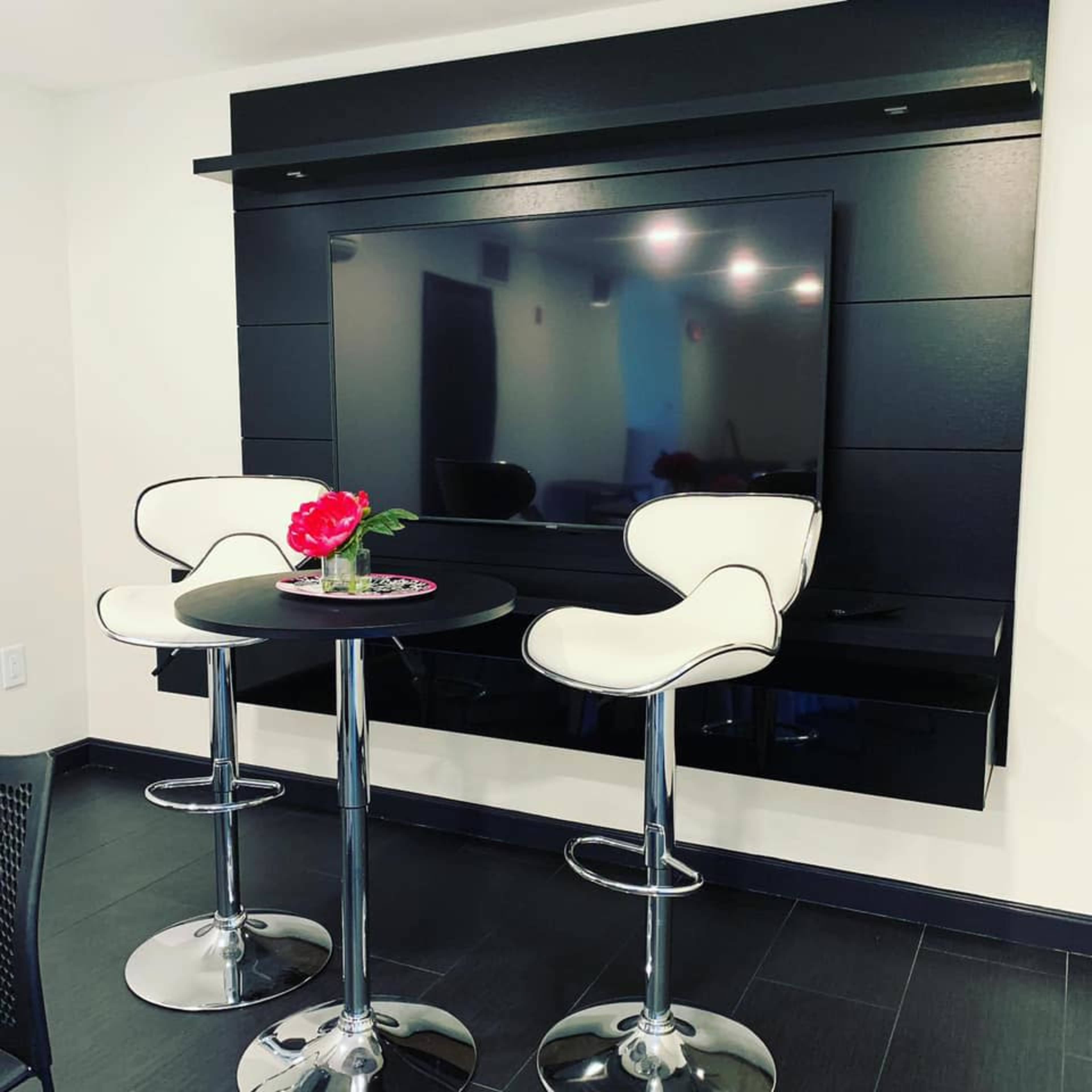 A modern black wall features a large flat-screen TV above a round table with two white bar stools and a flower centerpiece.